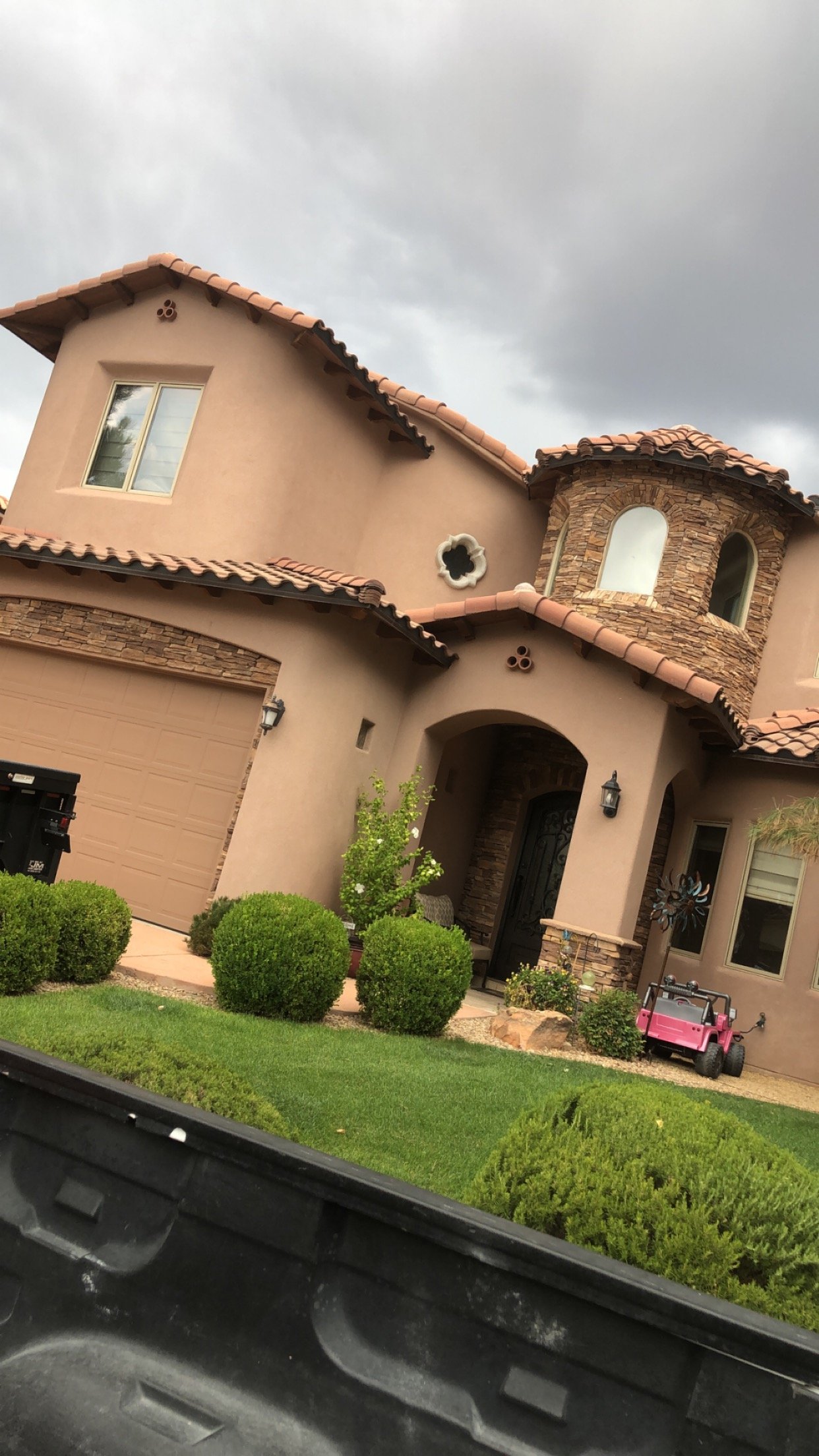 Front view of a house with a stucco exterior, tiled roof, and a small front yard with trimmed bushes and a pink toy car. The sky is overcast.