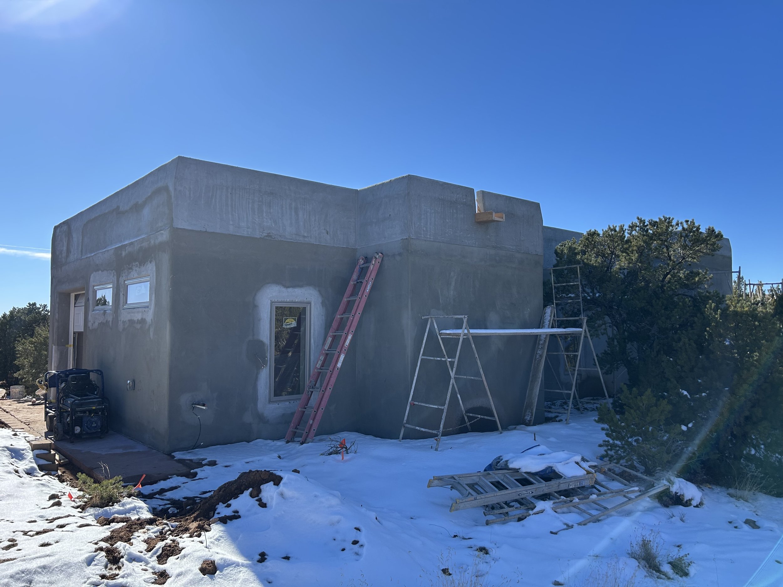 A two-story house under construction with gray exterior walls, surrounded by snow, construction ladders, and scaffolding, with trees and a clear blue sky in the background.