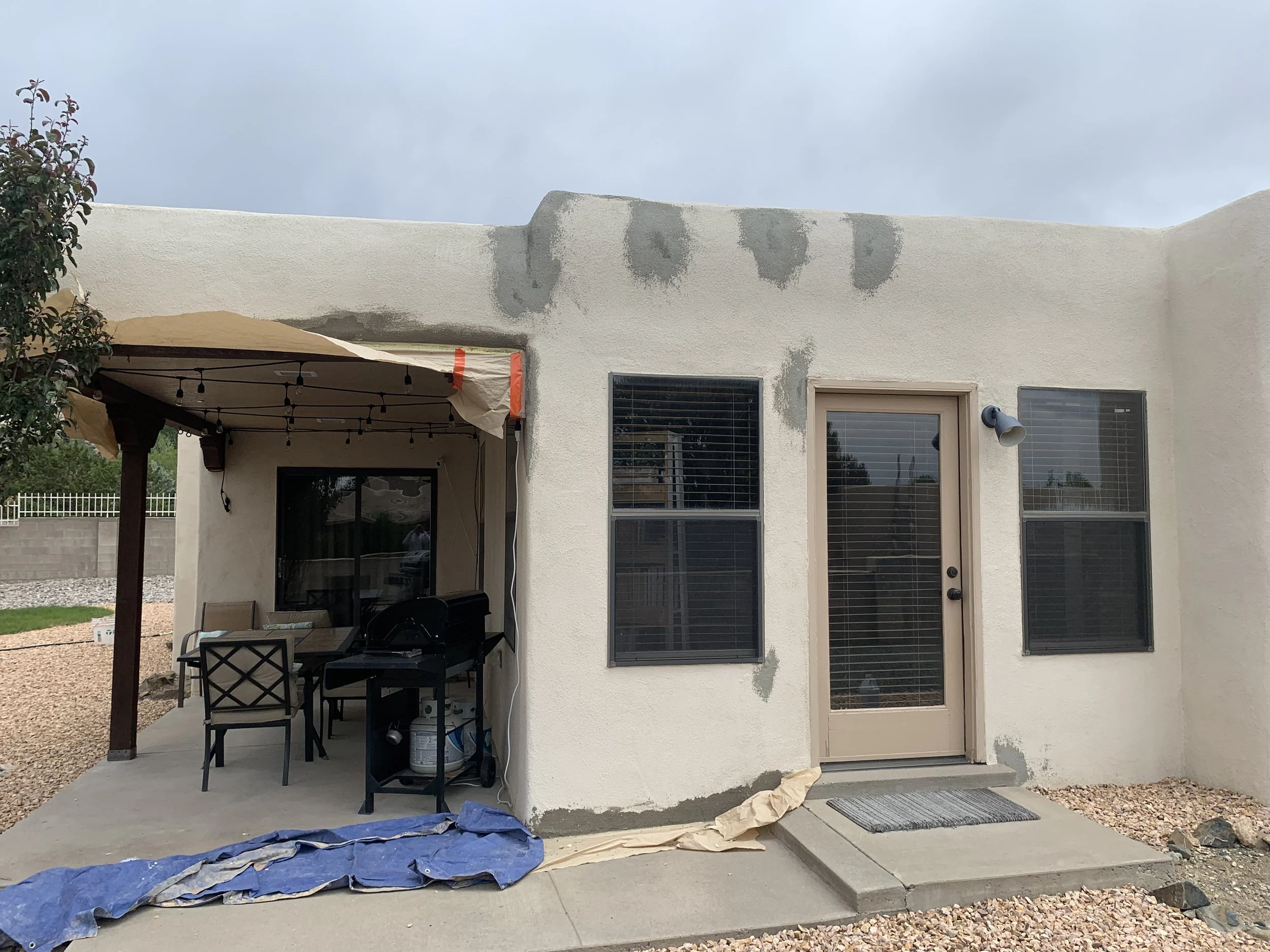 The back exterior wall of a house with patches of fresh grey stucco repair. A small patio area with a table, chairs, and a barbecue grill is visible under a beige canopy. A plastic tarp and brown paper are on the ground, and a door with a storm door and two windows are on the right side of the house.