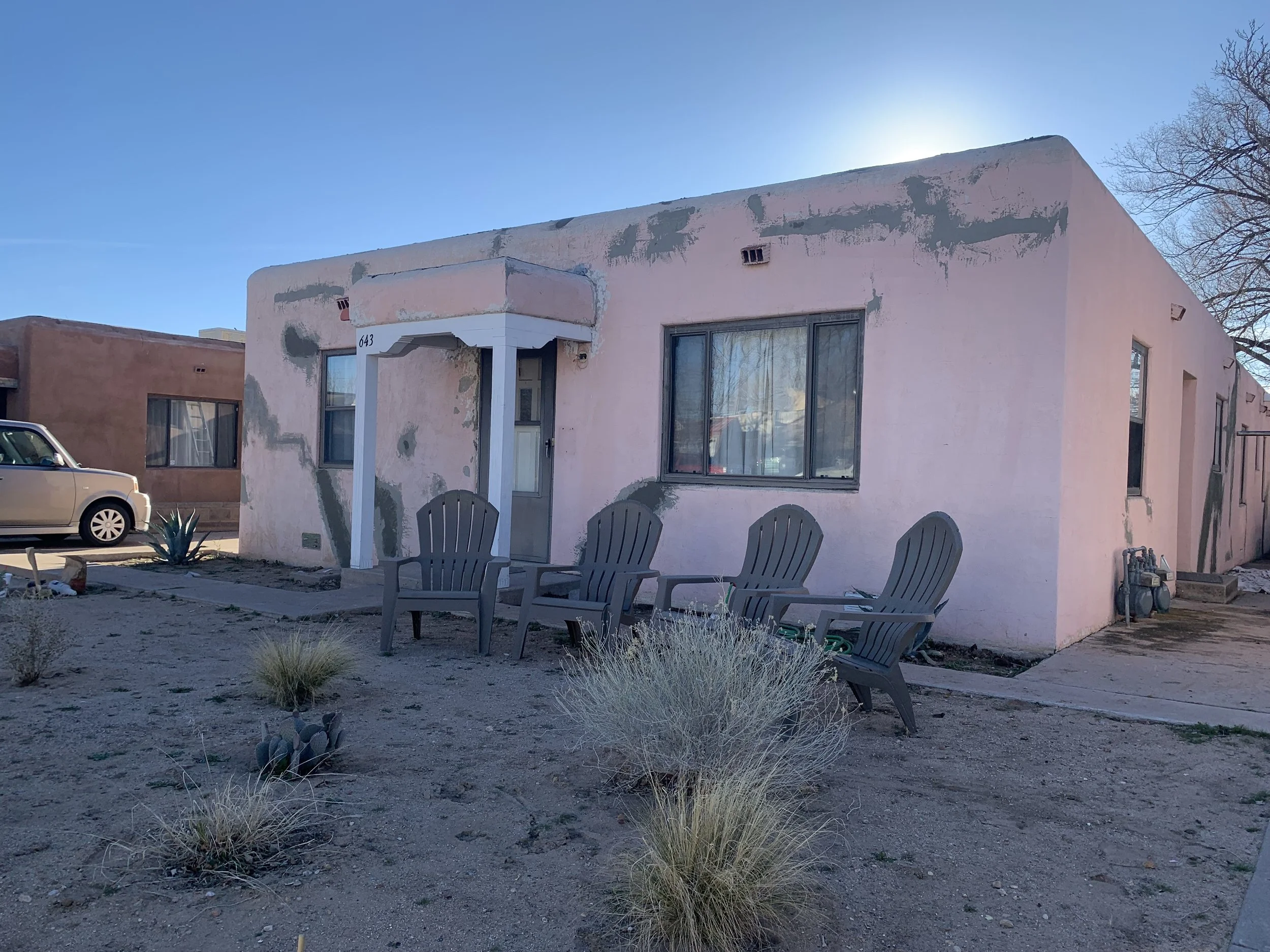 A pink stucco house with a flat roof showing patches of peeling paint and weathering. There are large windows and a small porch with two chairs outside. A car is parked on the street nearby, and desert plants grow in the front yard. The sky is clear and sunny.