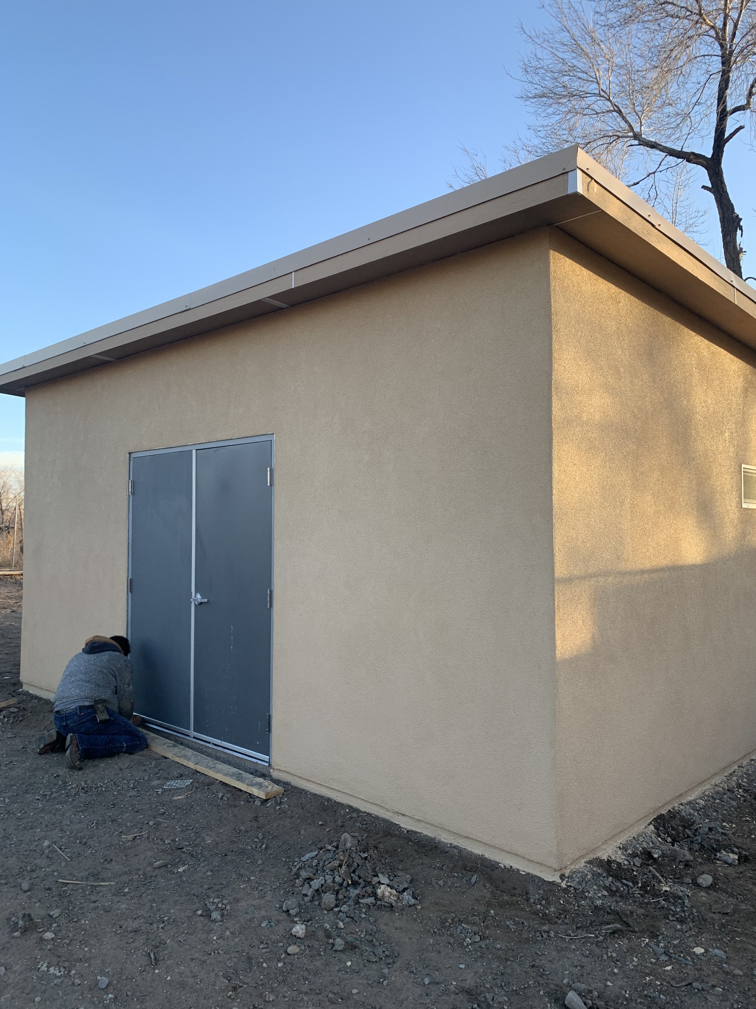 A beige building with a blue door, two workers installing a metal frame at the bottom of the building, and a tree with no leaves in the background.