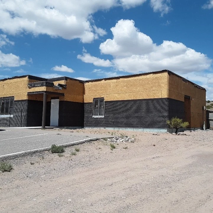 A building under construction with partially finished walls, a parking lot in the foreground, and a partly cloudy sky above.
