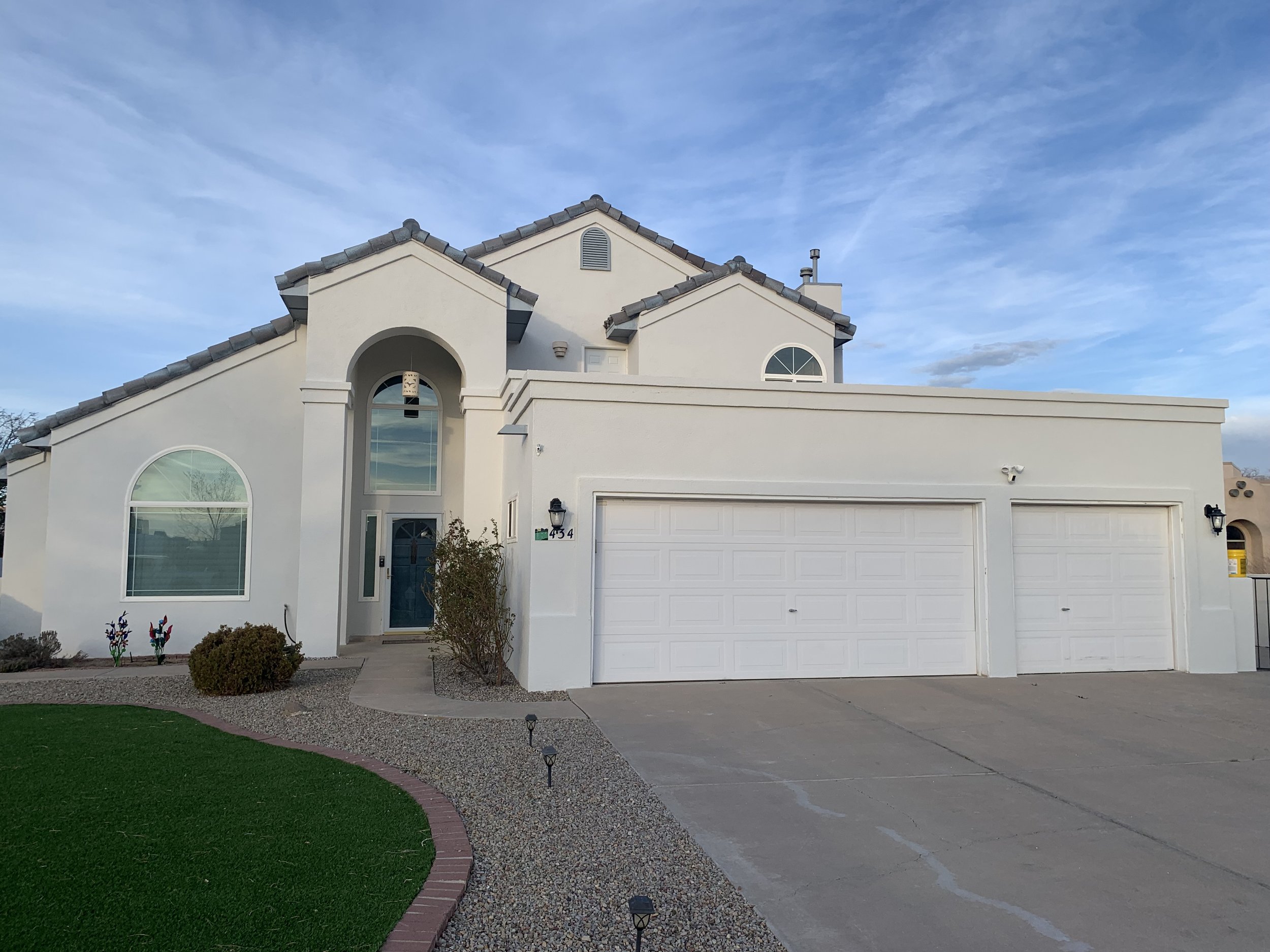 A white two-story house with arched windows and a garage door, front yard with a curved gravel pathway, green lawn, and some plants, under a blue sky with some clouds.
