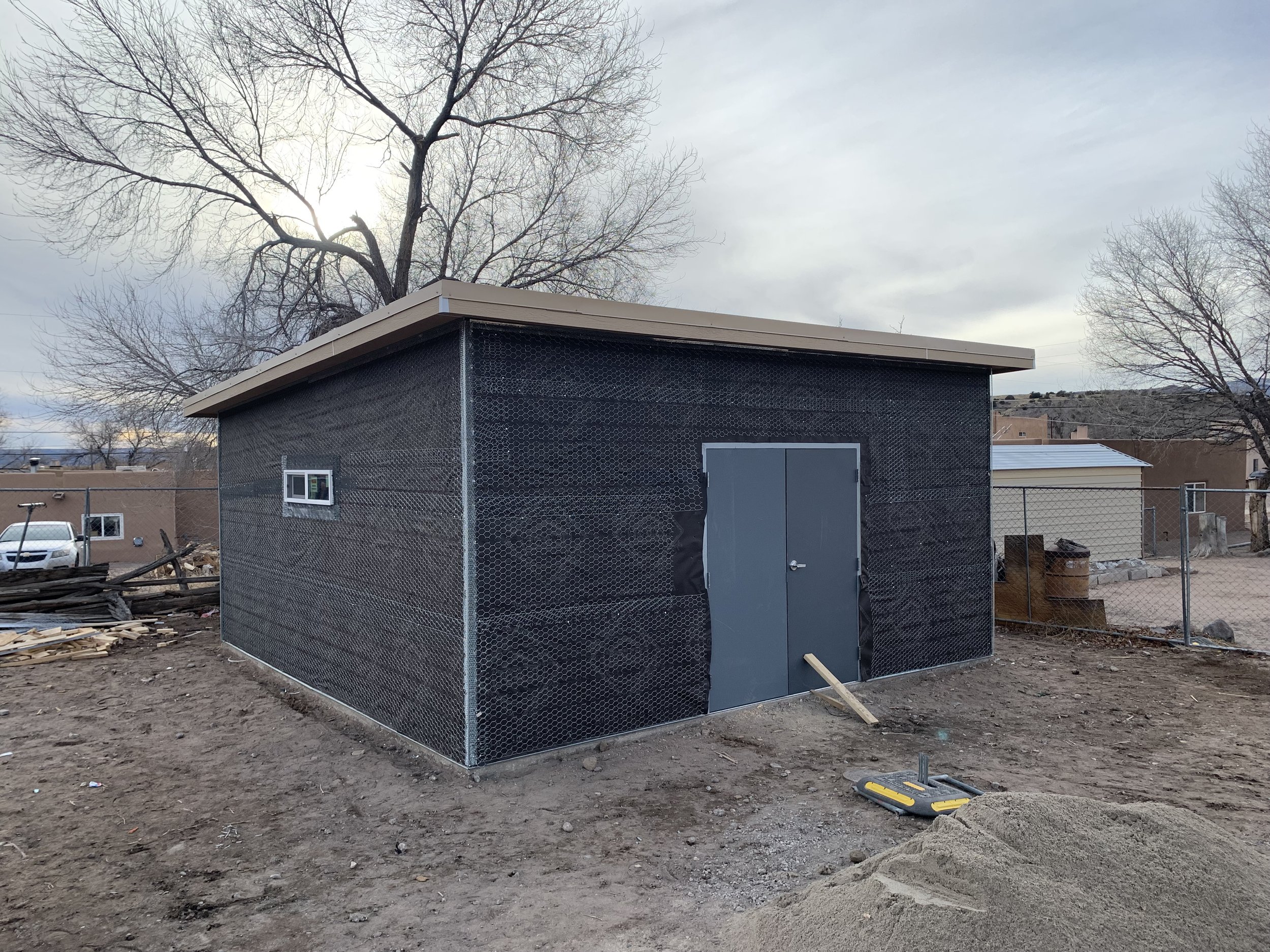 Small black building with a gray door and window, surrounded by a chain-link fence, on a barren dirt lot with some construction materials and tools, under a cloudy sky with leafless trees in the background.