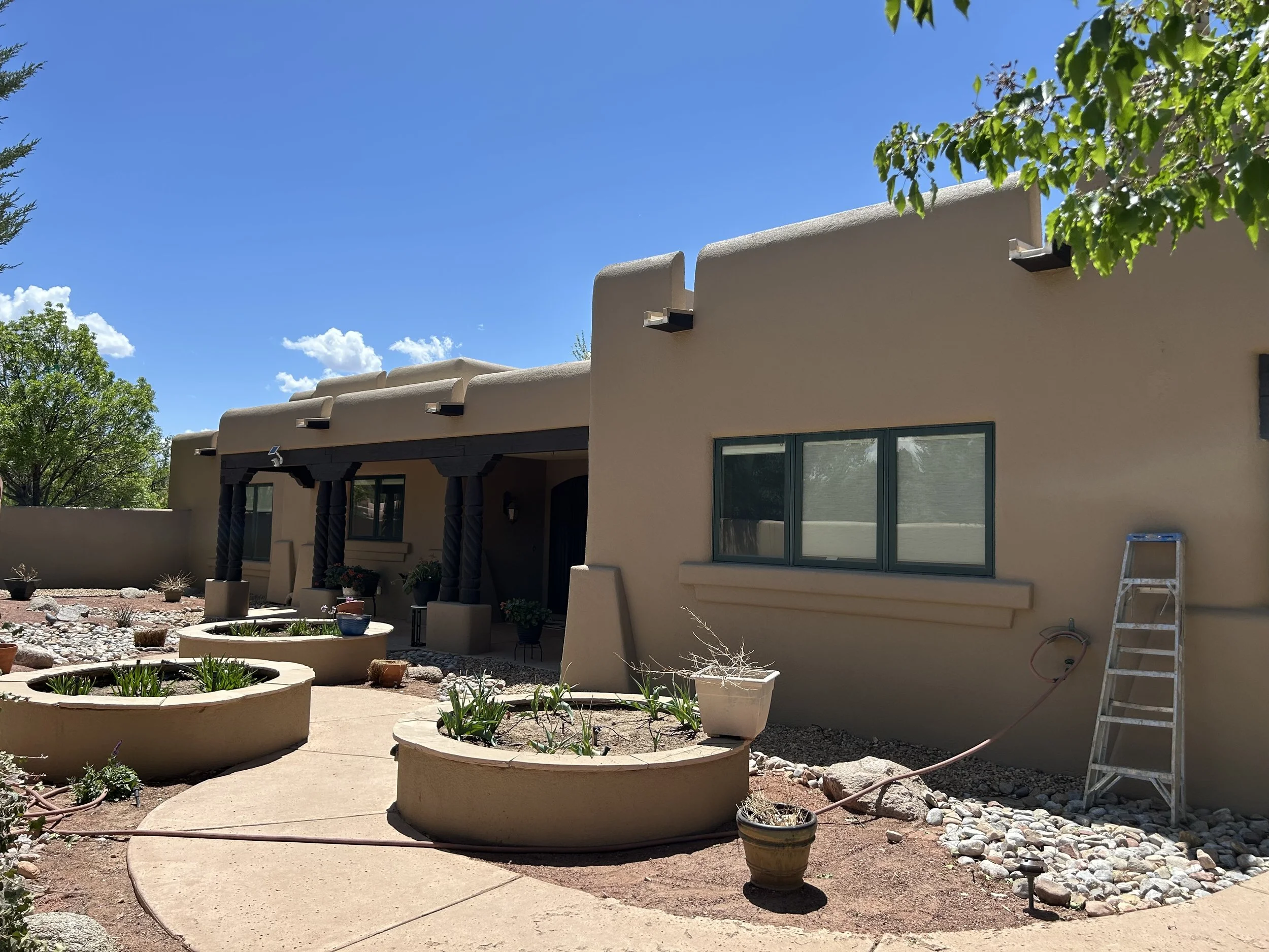 The image shows the backyard of a house with beige stucco walls and large windows. The backyard features a patio area with a pergola and potted plants. There are curved concrete planters with plants, a garden with rocks, and a ladder leaning against the house. Green trees and a blue sky with some clouds are visible in the background.