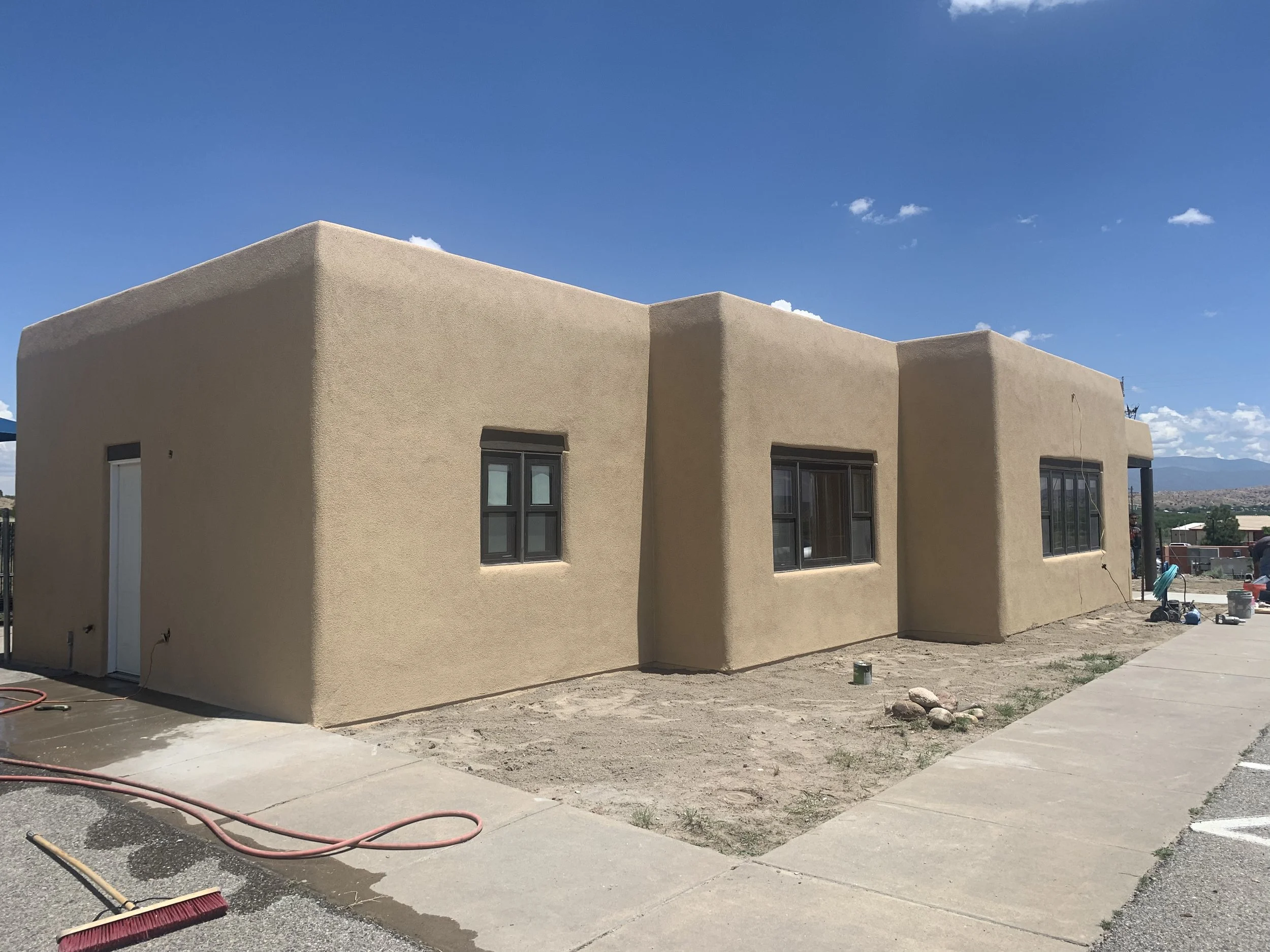 A beige stucco building under construction with a flat roof, surrounded by unfinished concrete sidewalk and ground. Construction tools and supplies are visible, with a mostly clear blue sky overhead.