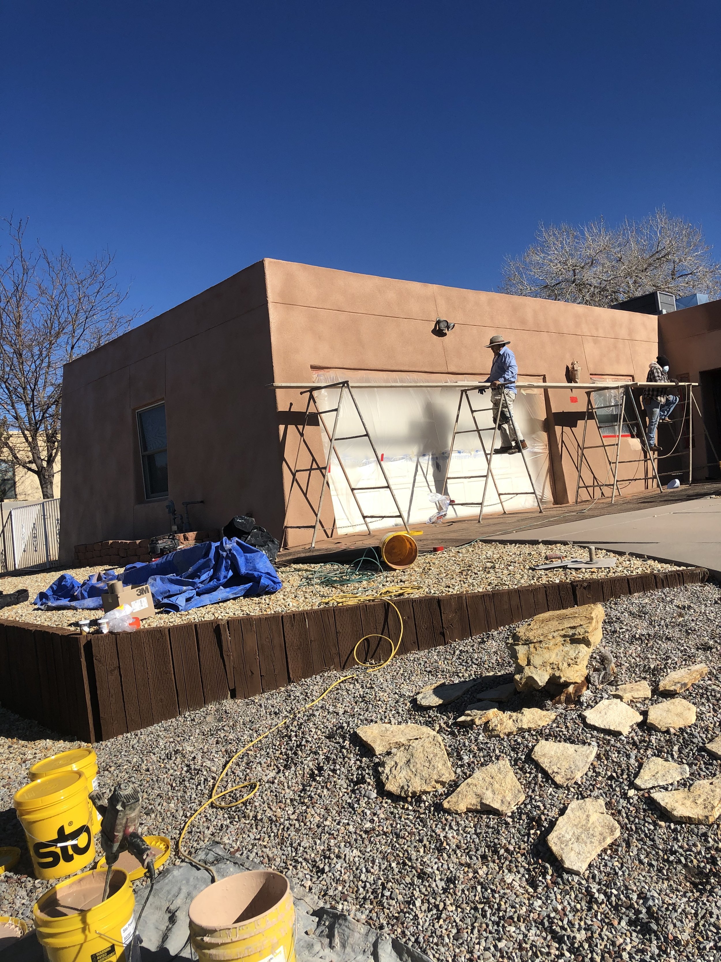 Workers on scaffolding applying a coat of paint or plaster to the exterior wall of a building. The building has a stucco finish, and the workers are dressed in casual clothing with hats. Construction tools and supplies are scattered on the ground.