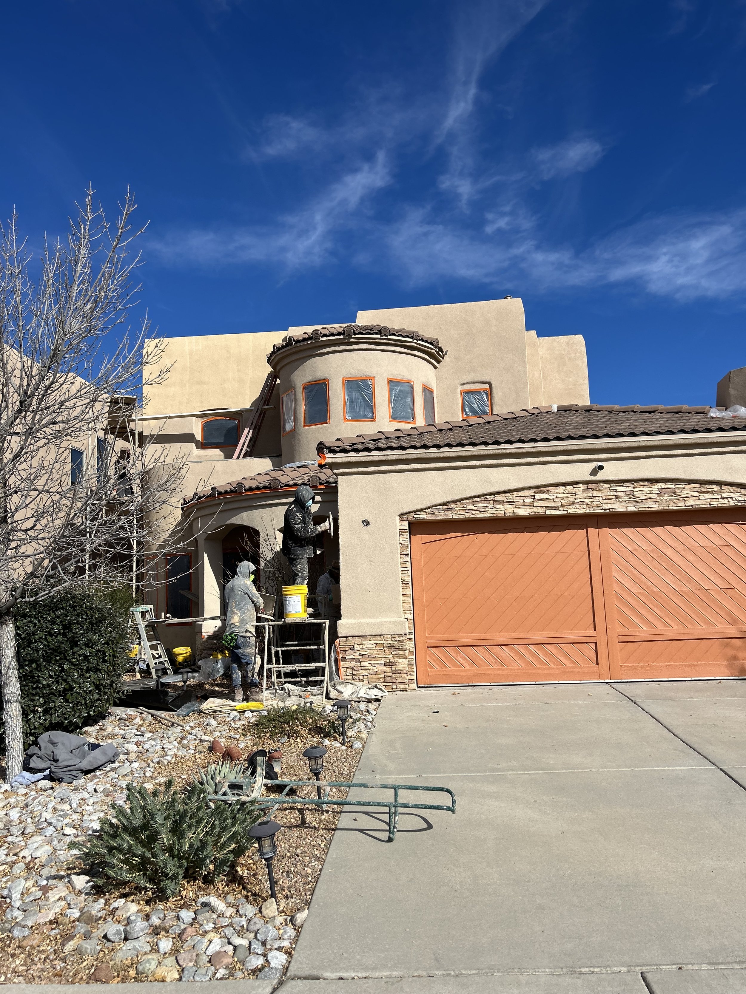 Workers in winter clothing working on the exterior of a house, painting or remodeling, with a light-colored stucco finish, a decorative rounded window area, and an orange garage door, under a clear blue sky with some clouds.