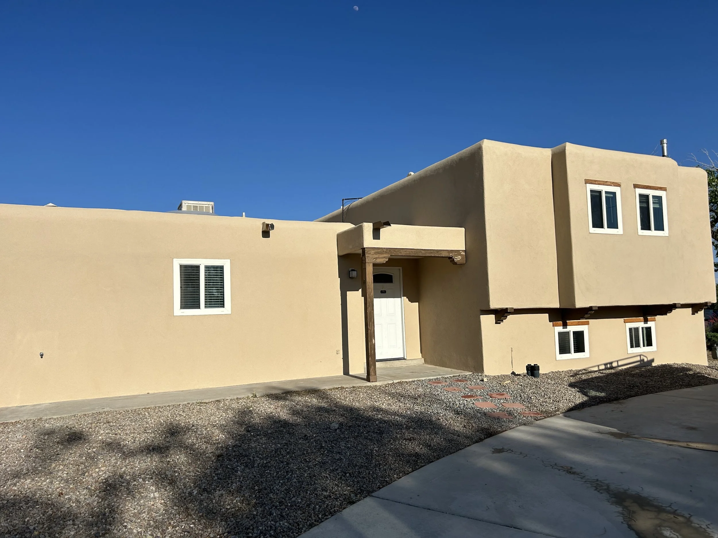 Front view of a modern beige stucco house with several windows, a white door, and a flat roof, set against a clear blue sky.