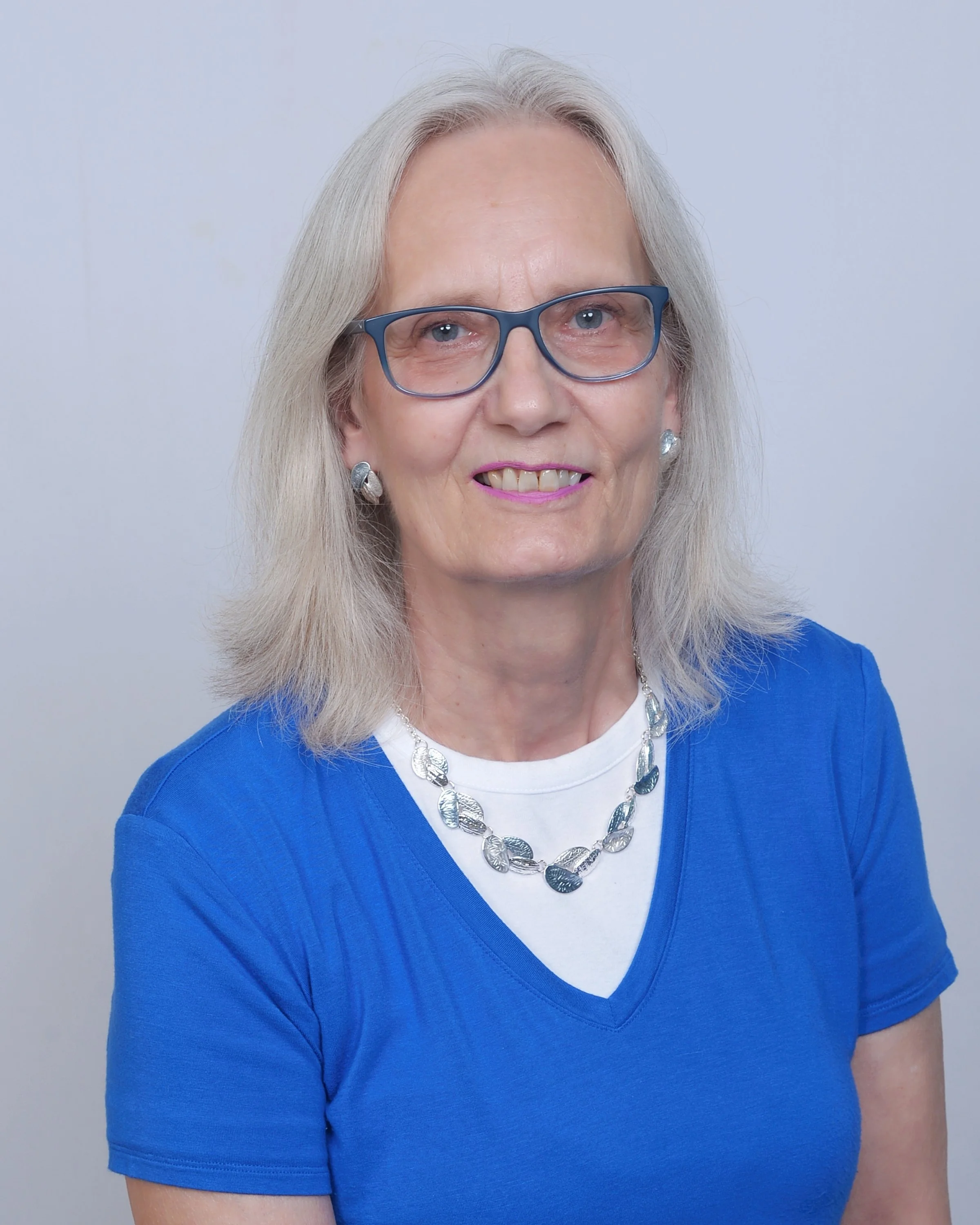 Close-up of a smiling older woman with gray hair, wearing glasses, a white shirt, blue cardigan, and a silver necklace against a plain background.