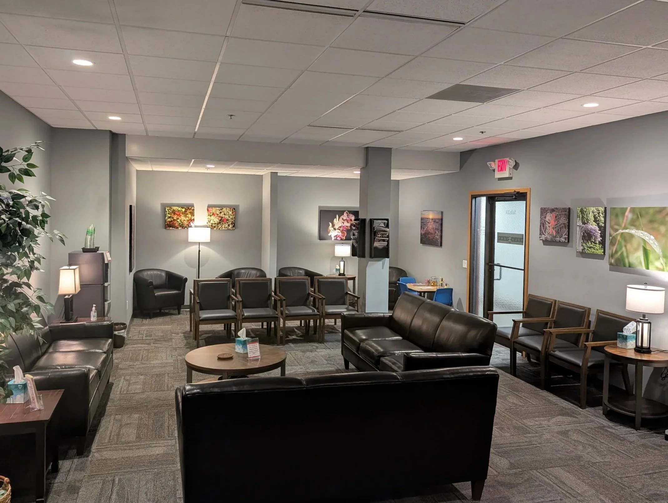 Indoor waiting area or lobby with black leather sofas, chairs, small tables, framed floral artwork on light gray walls, potted plant on the left, floor lamps, and a glass door with an exit sign above.