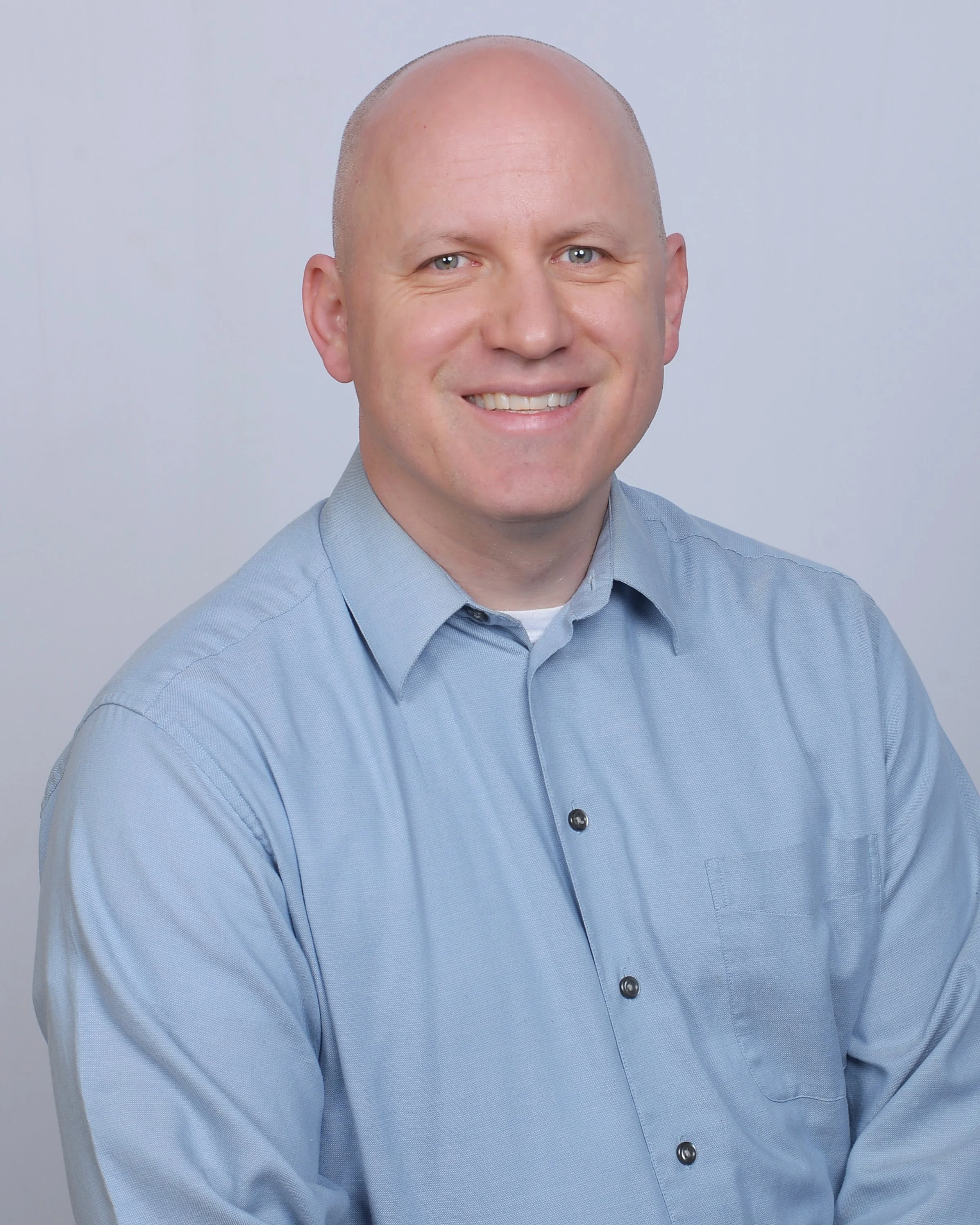 Portrait of a smiling man wearing a light blue dress shirt, with a bald head, light skin, and blue eyes, against a plain light background.