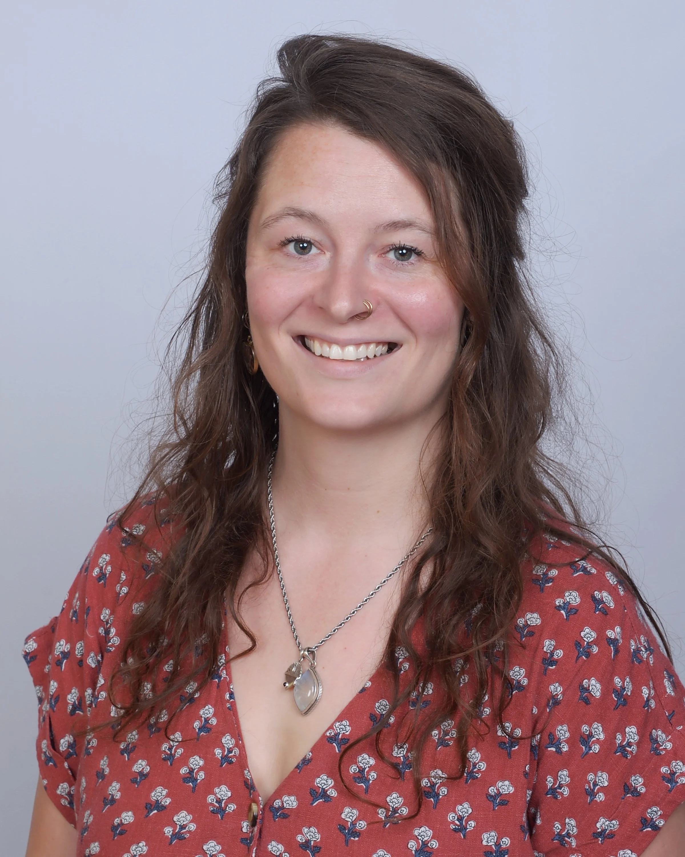A woman with long, wavy brown hair styled with volume, wearing a red blouse with a floral pattern, silver necklaces, hoop earrings, and a nose ring, smiling against a plain white background.