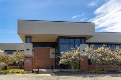 Modern office building with brick and glass exterior, trees in front, under a partly cloudy sky.