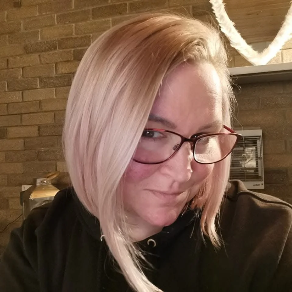 A woman with shoulder-length blonde hair, pink highlights, and glasses smiling at the camera. She is in a room with a brick wall background.