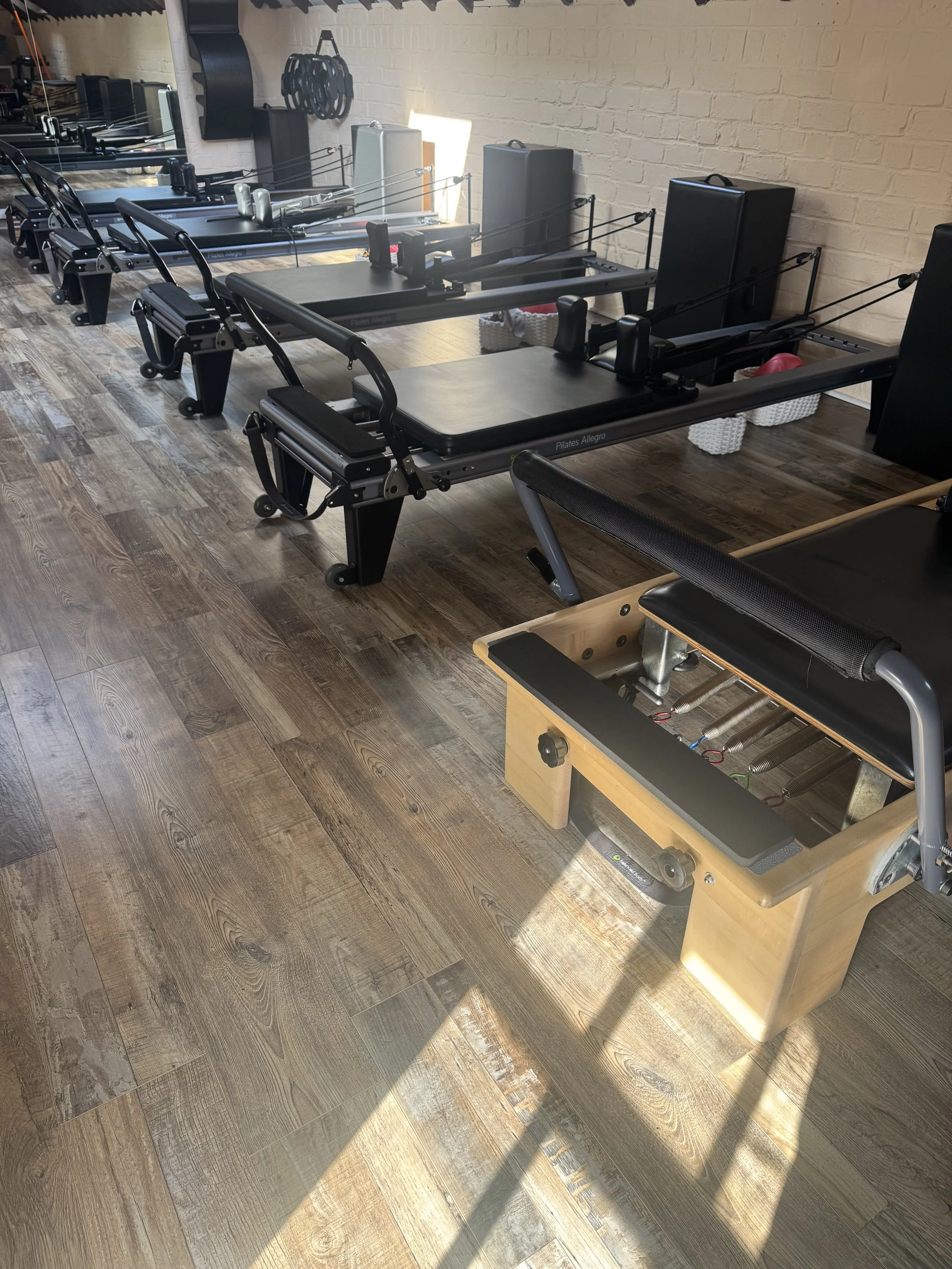 A Pilates studio with multiple Pilates reformer machines arranged in a row on a wooden floor, with a brick wall in the background.
