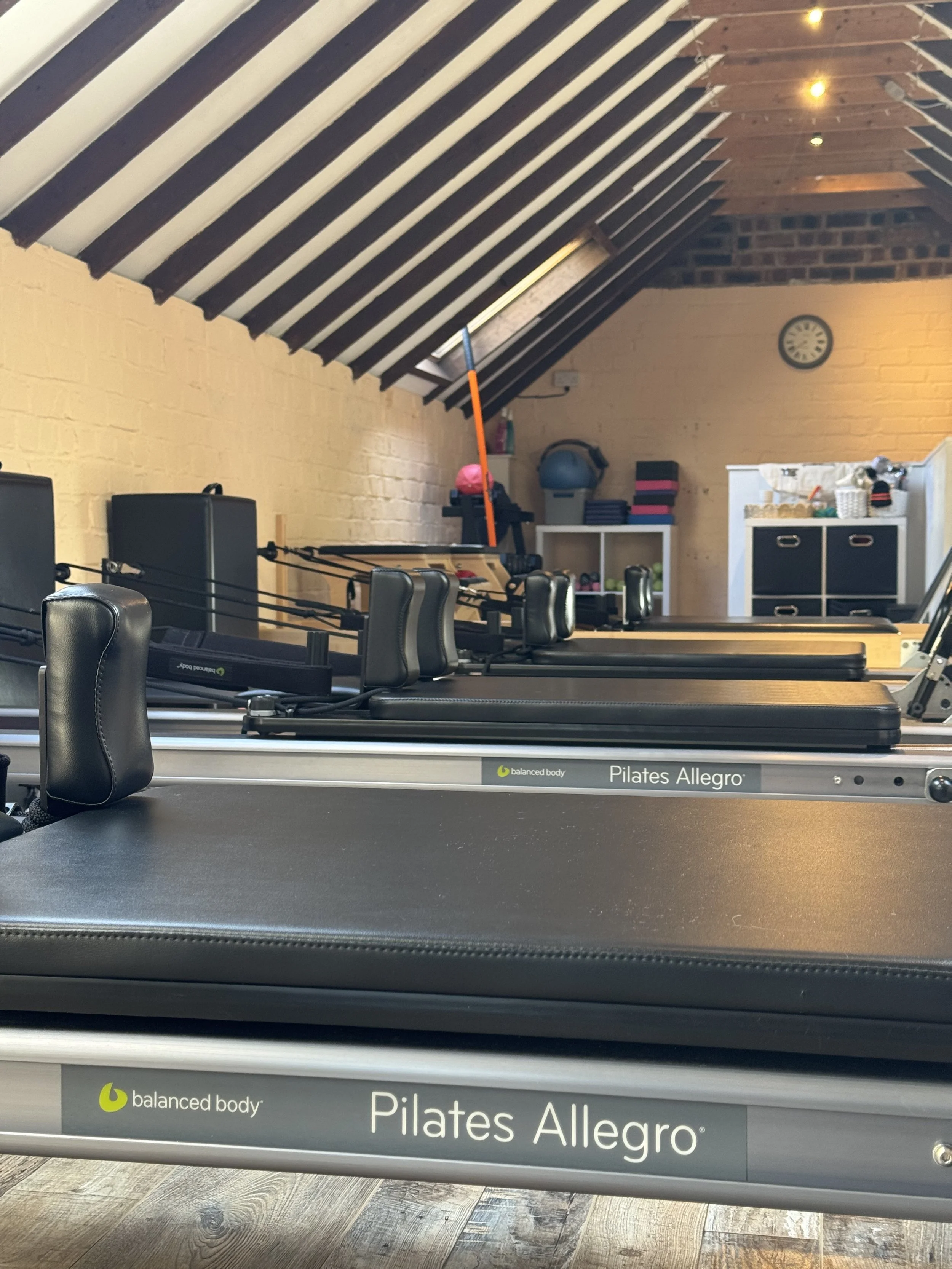A Pilates Allegro reformer machine in a fitness room with yellow brick walls and wooden ceiling beams. The room includes workout equipment like kettlebells, a large exercise ball, and shelves with various fitness accessories.