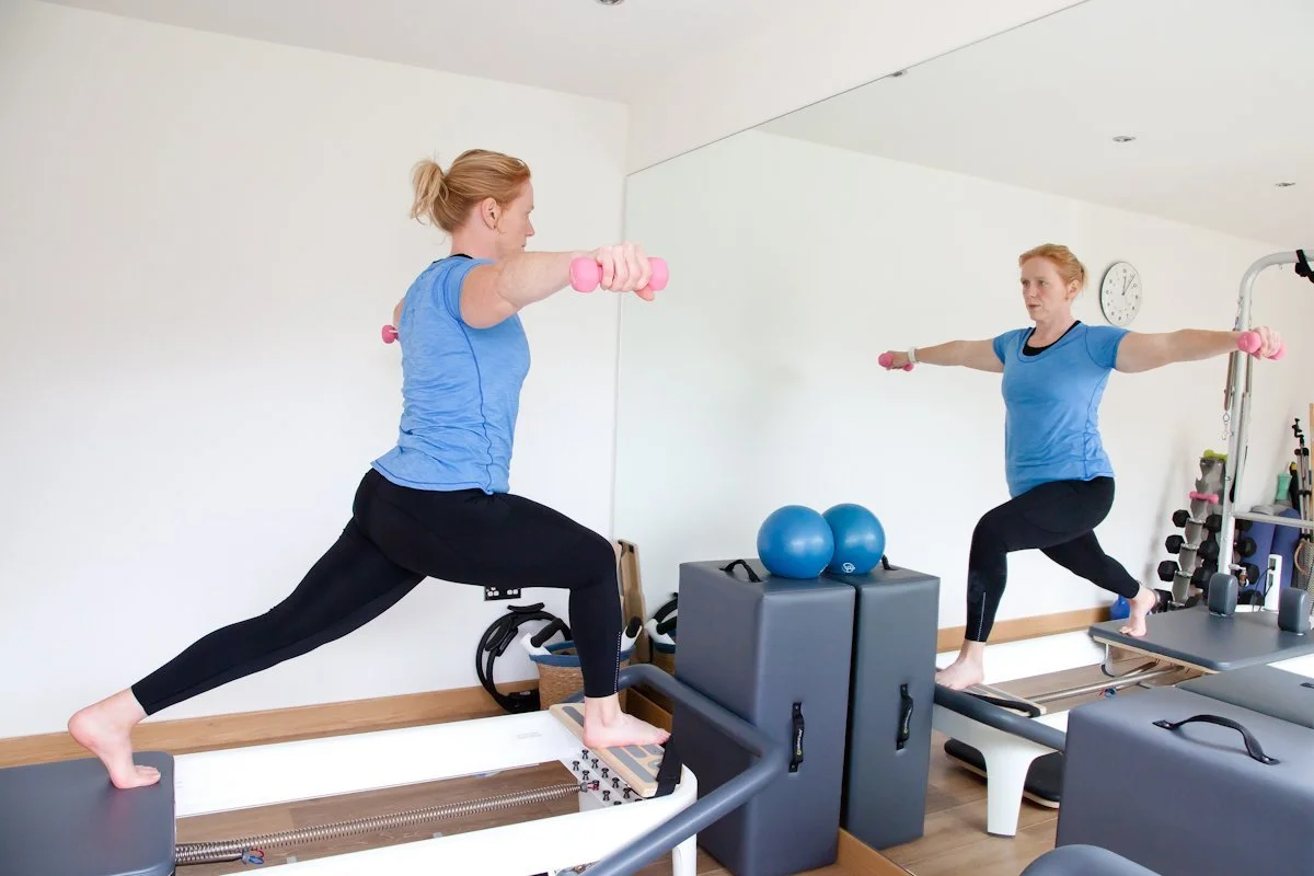 A woman is exercising on reformer machines in a gym, holding pink dumbbells with her arms extended outward and one foot on the machine, with a mirror reflecting her image.