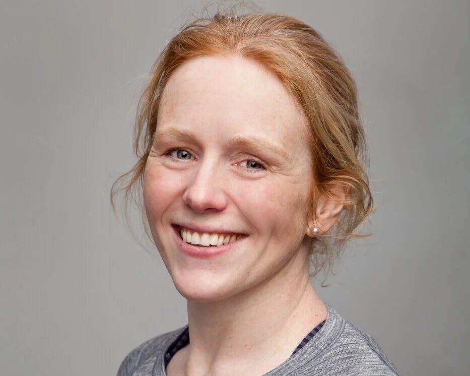 A young woman with red hair, fair skin, and wearing a gray top, smiling against a plain gray background.