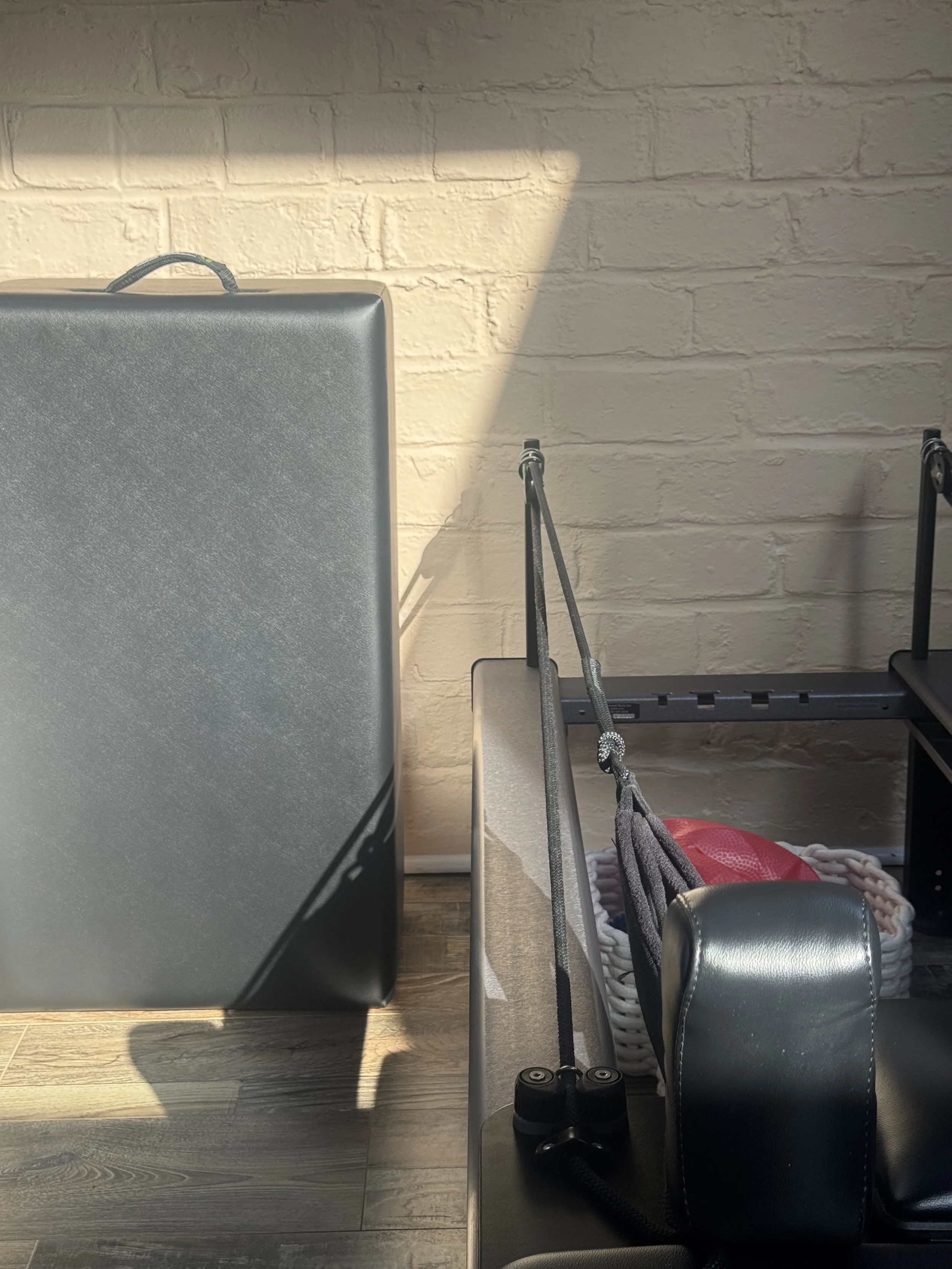 Indoor scene with exercise equipment, including a black treadmill, a chair, a basket with ropes, and a gray padded chair, against a beige brick wall and wooden floor.