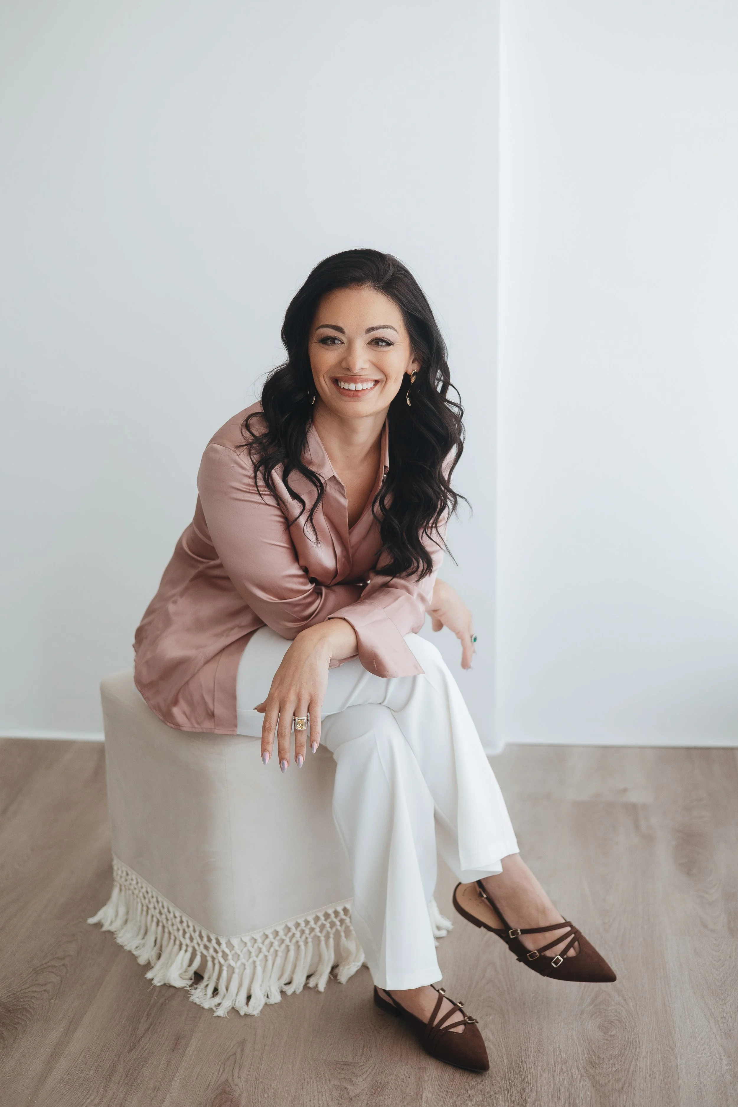 A woman with long dark wavy hair, wearing a pink blouse and white pants, sitting on a white woven stool with fringe, in front of a plain light-colored wall. She is smiling, showing her teeth, and wearing rings and earrings.