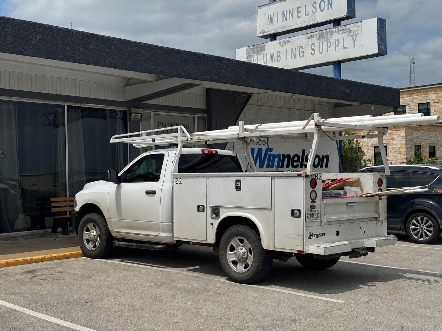 White utility truck parked in front of a plumbing supply store, with ladders and tools loaded on the truck's rack.