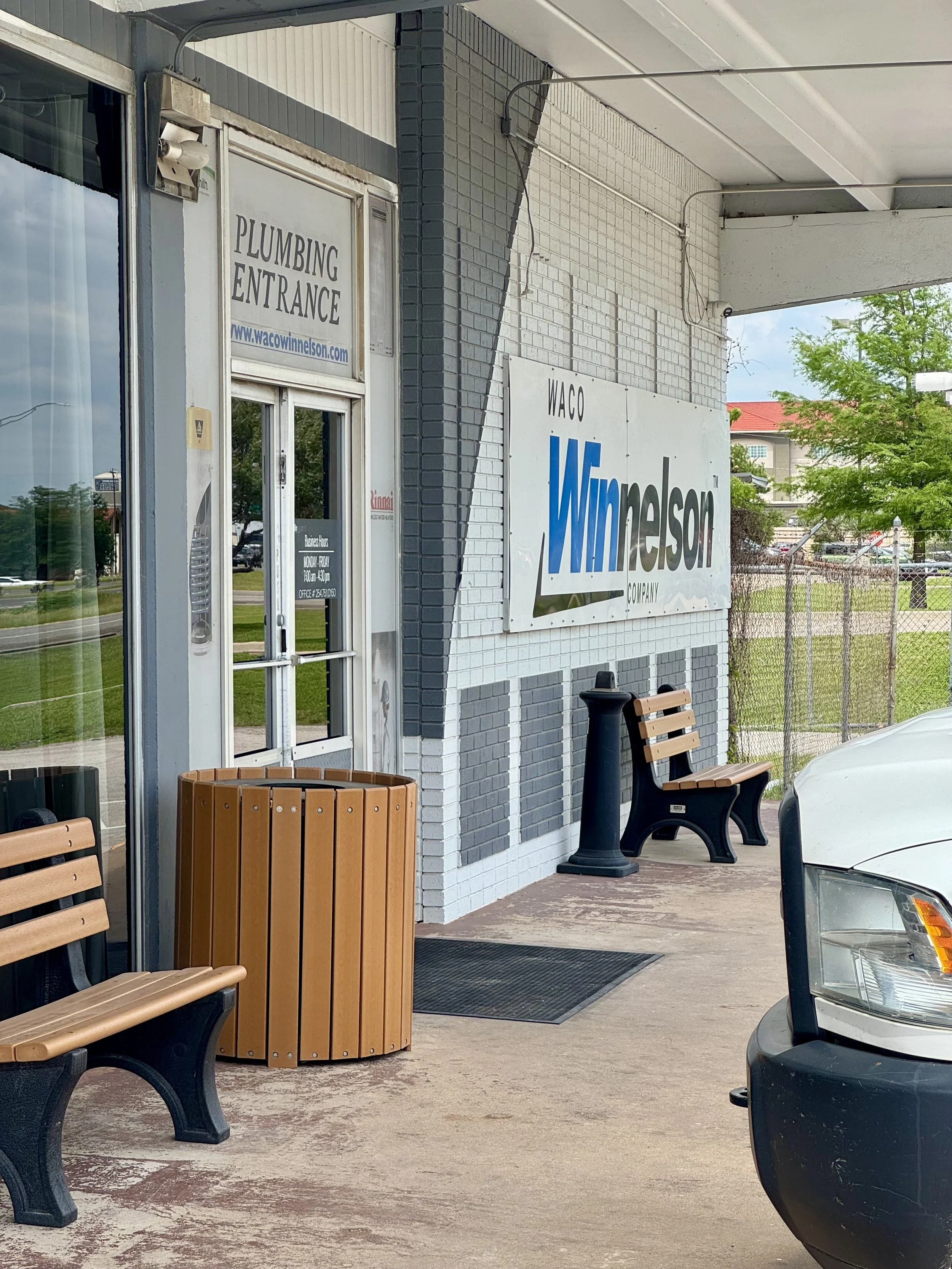 Exterior of a commercial building with signage for Waco Windsor company and plumbing entrance. There are wooden benches, a trash can, and a white car parked nearby, with trees and a grassy area in the background.