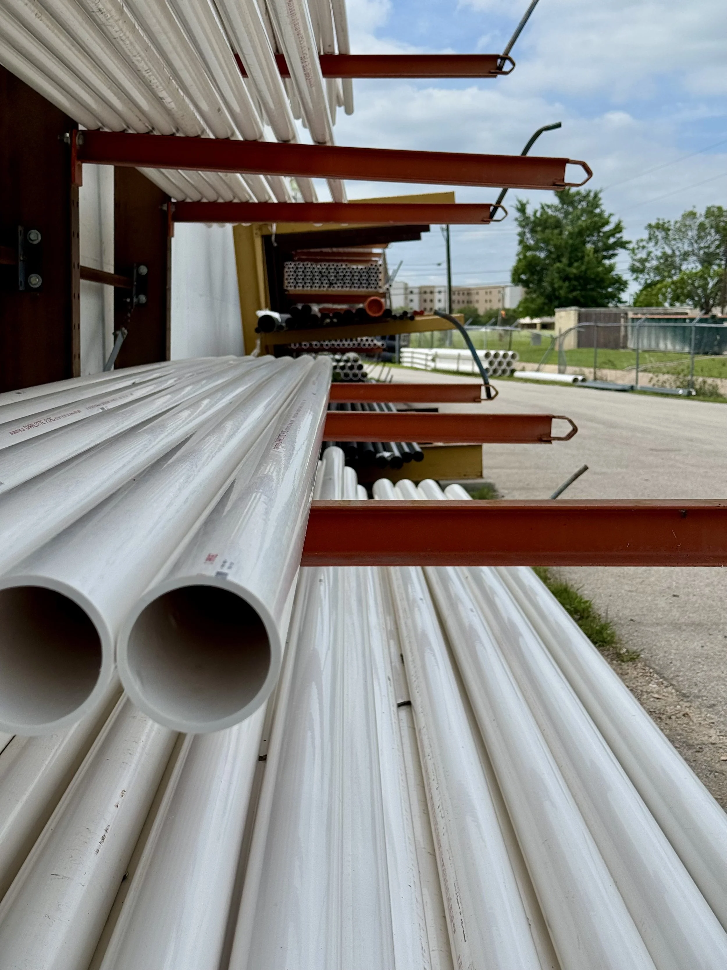 Rows of white pipes and orange metal racks outdoors with a grassy area, trees, a fence, and buildings in the background under a cloudy sky.