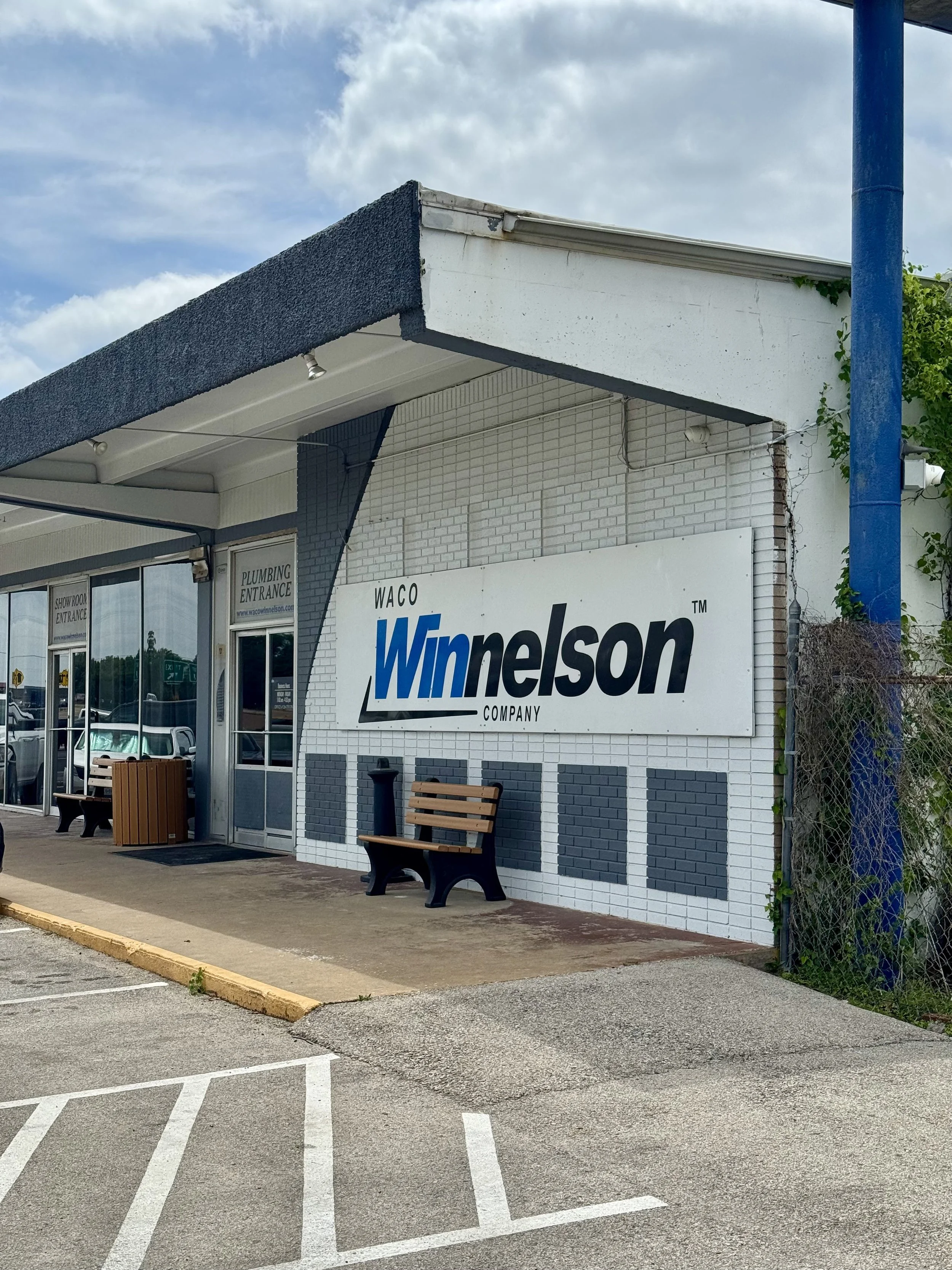 The exterior of a Winnelson plumbing supply store with a large sign, a blue pole, benches, a trash can, and a parking lot with marked spaces.