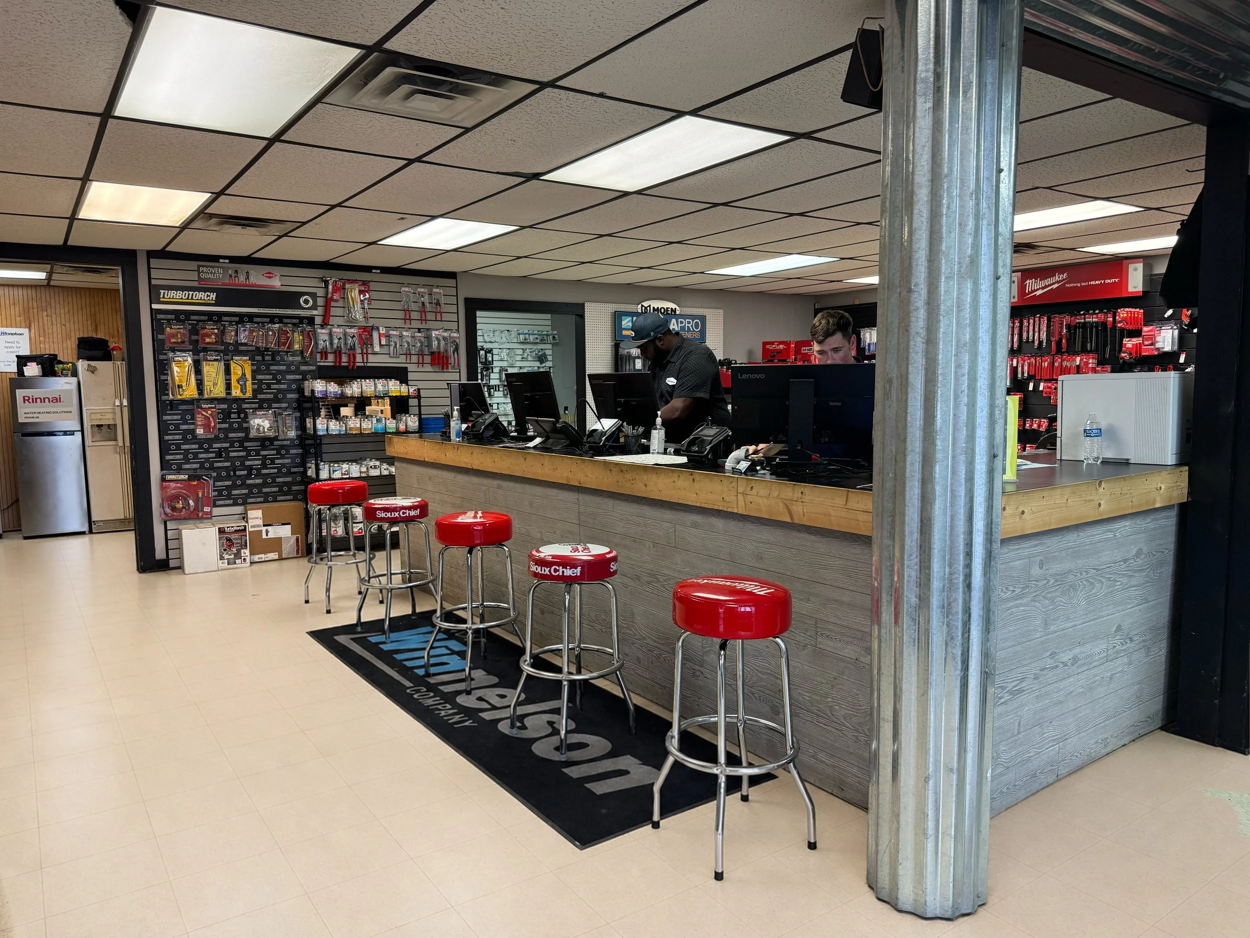 View of an automotive parts store checkout counter with employees working, surrounded by shelves of tools and supplies, with red stools in front of the counter.