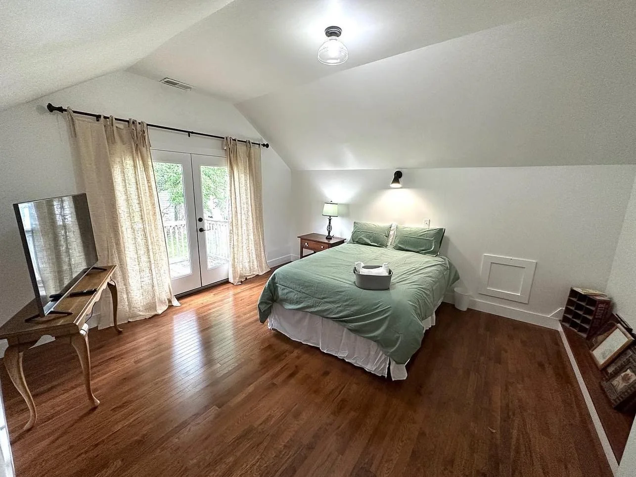 A bedroom with a bed, nightstand, lamp, flat-screen TV on a wooden table, and glass doors with curtains leading to an outside space. The room has hardwood flooring and sloped ceilings.
