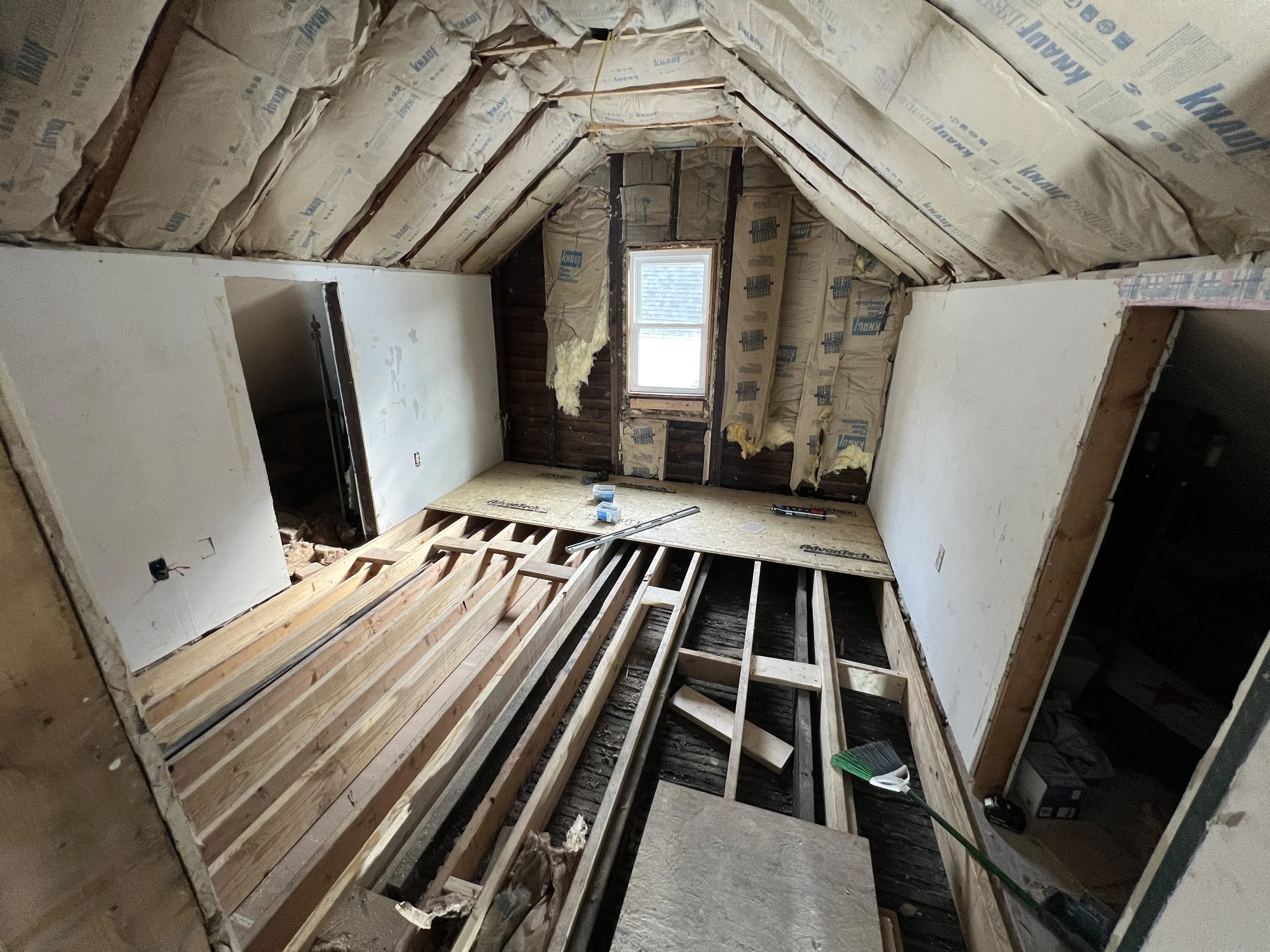 Attic undergoing renovation with exposed wooden floor joists, insulation, and drywall walls. A small window at the far end of the space.