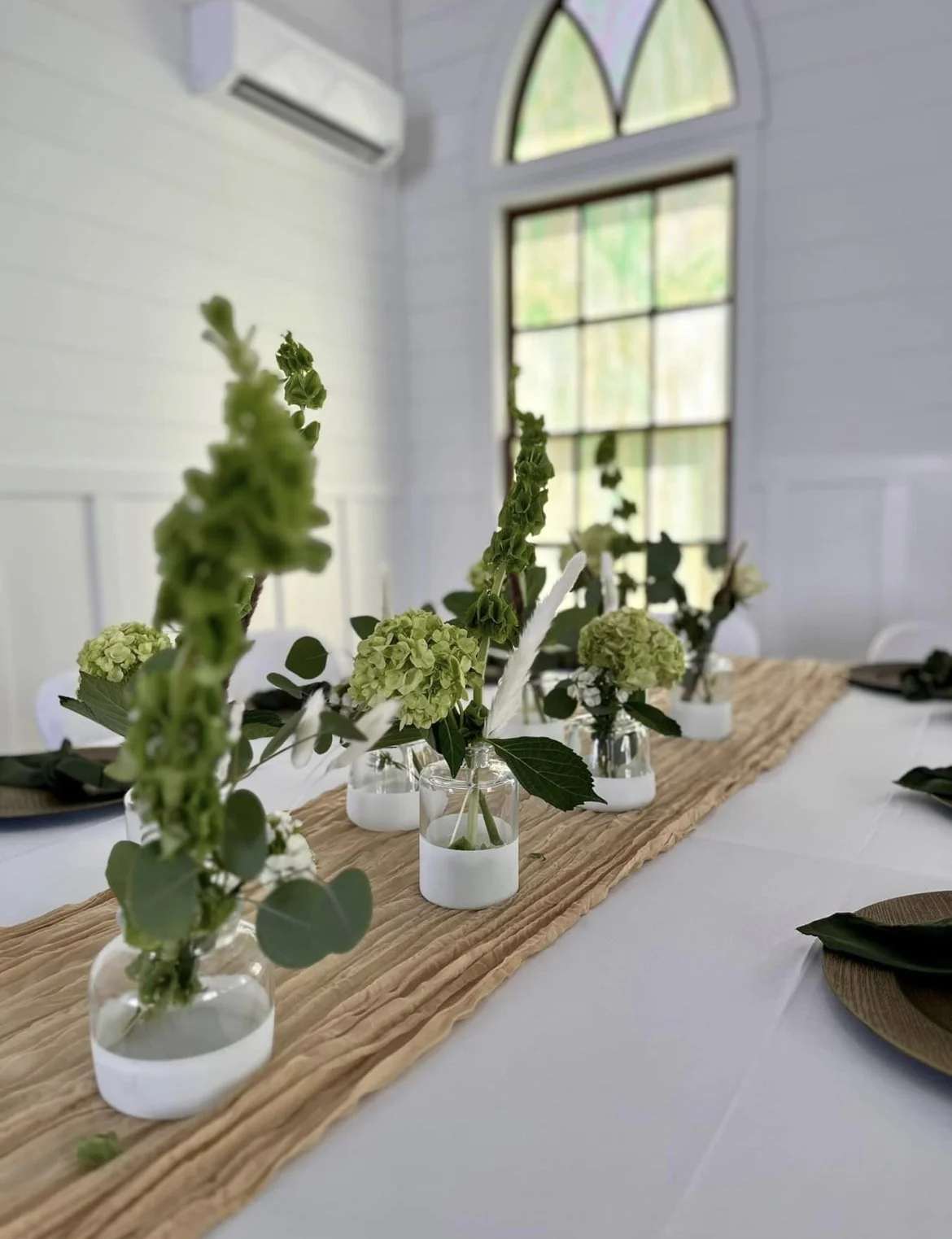 Table centerpiece with white and green flowers in small glass vases on a beige table runner in a bright room with large windows.