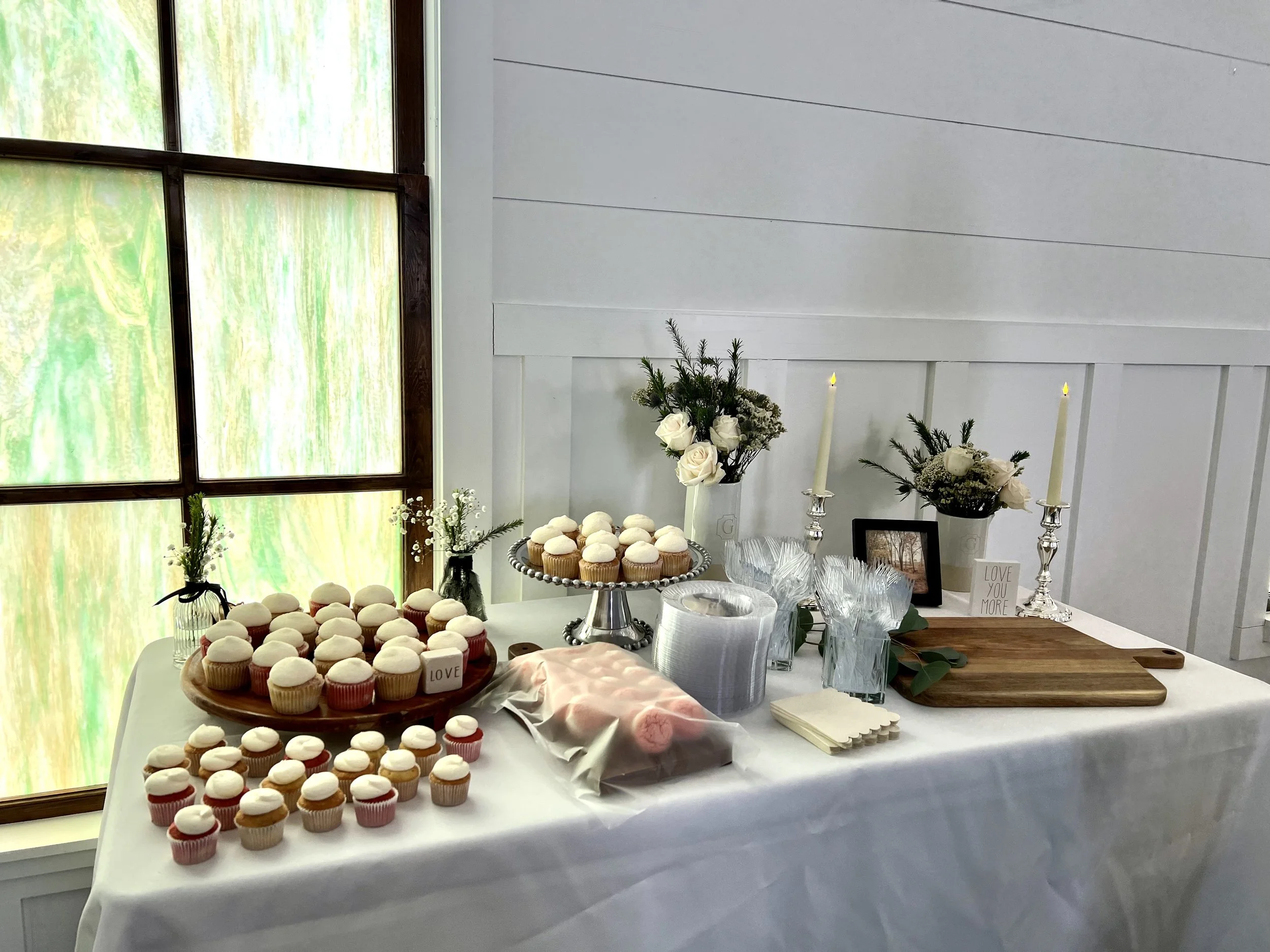 Table decorated with white flowers, candles, cupcakes, and framed photo, set against white wall and window.