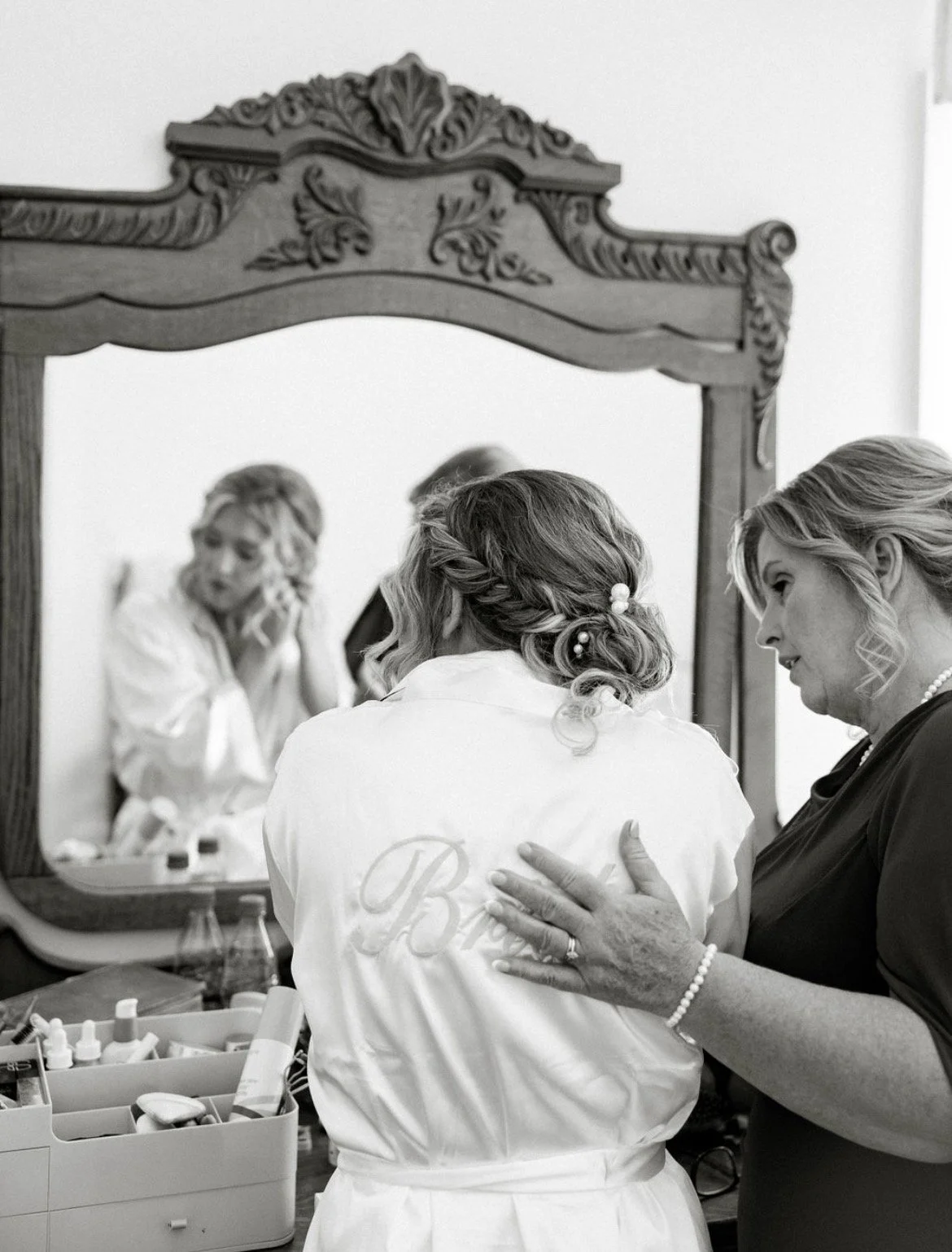 A woman getting ready, seen from behind, wearing a robe with a monogram, while an older woman assists her in a mirror reflection. The scene appears to be in a dressing room or preparing area.