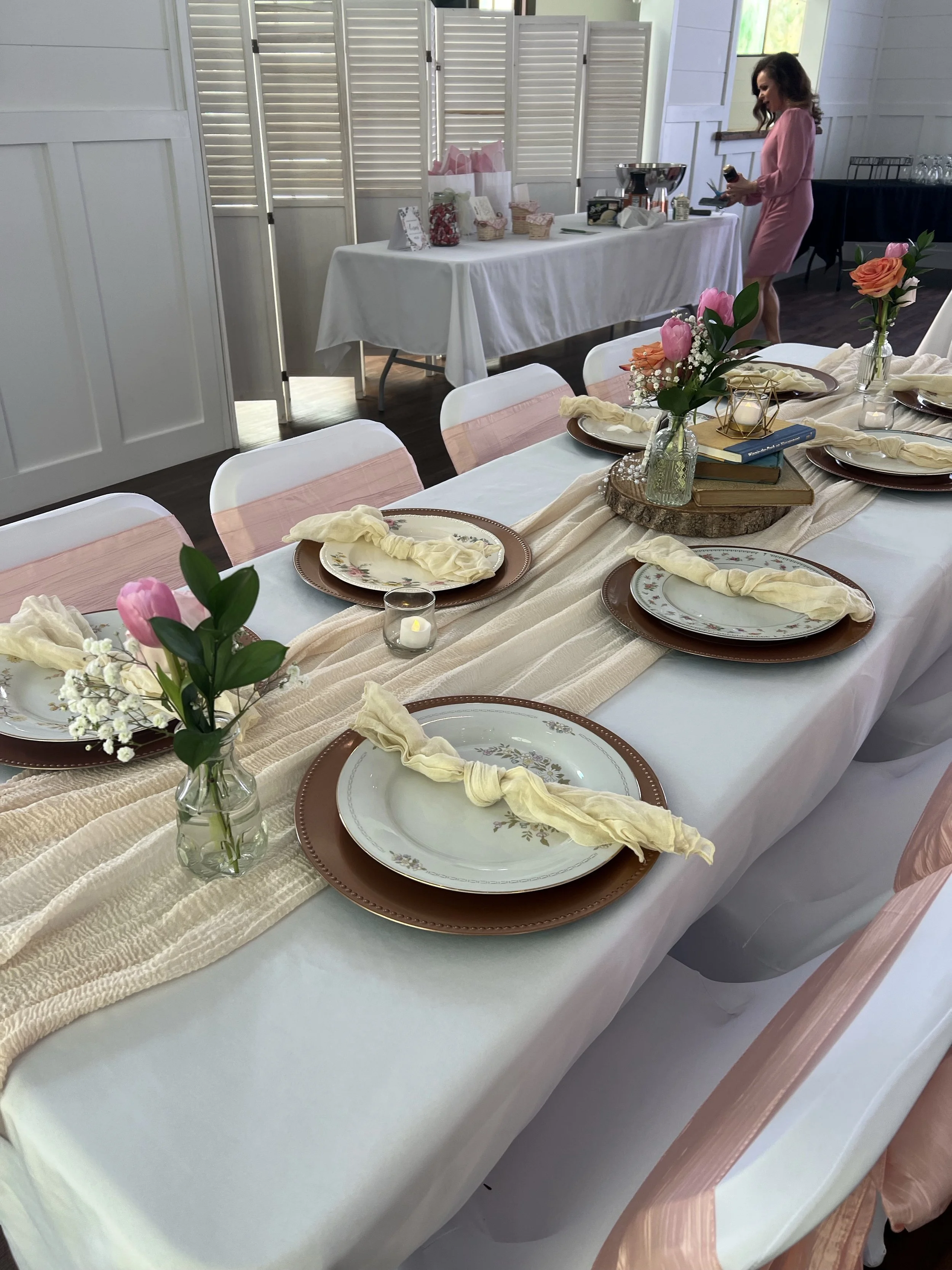 A table set for a celebration or event with white tablecloths, pink napkins, floral centerpieces, and place settings, with a woman in a pink dress standing in the background preparing food or serving.