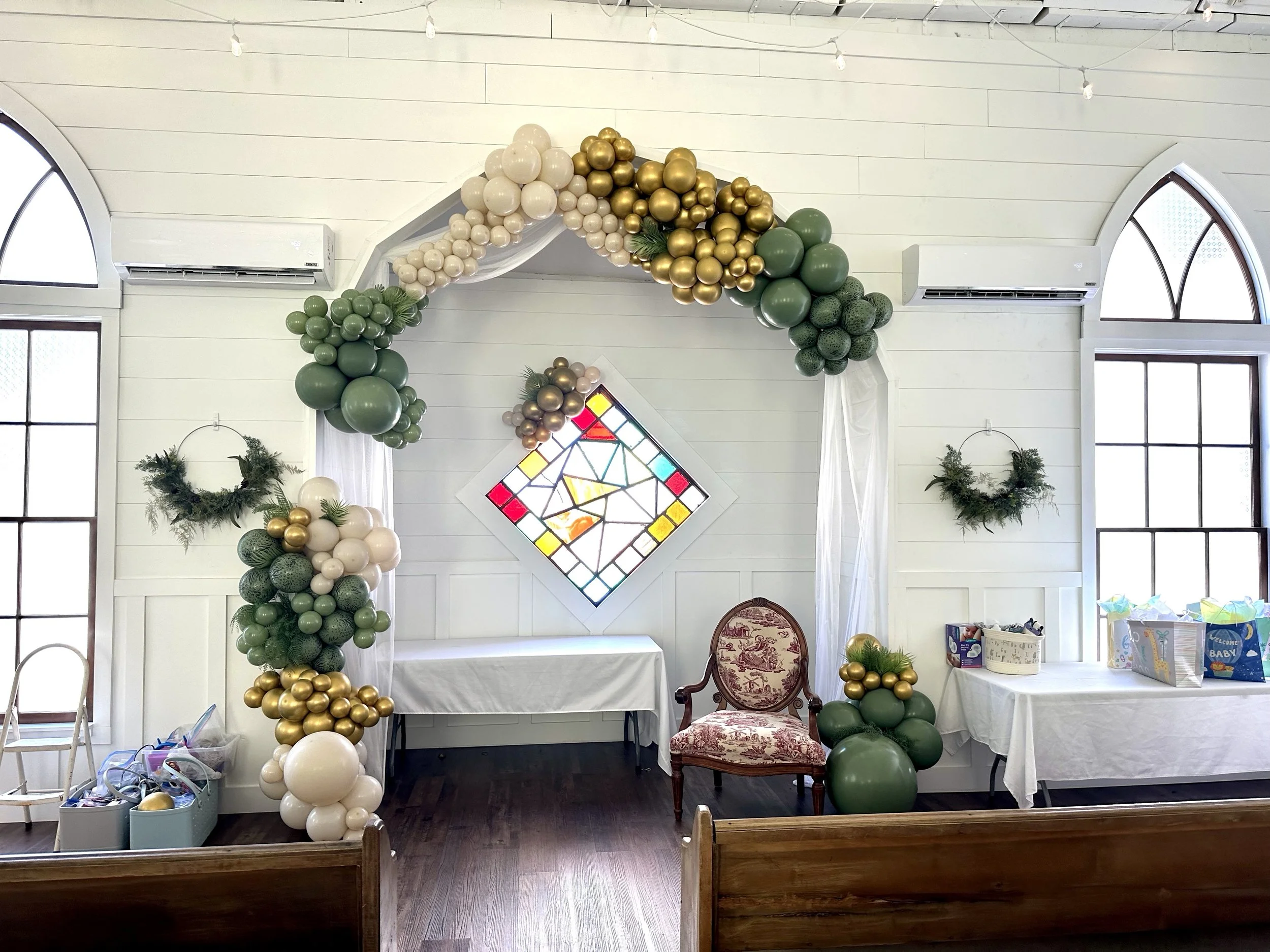 Decorated interior space for celebration with gold, cream, and green balloons arranged as an arch and clusters, inside a room with high windows, white walls, and stained glass window, with tables and chairs.
