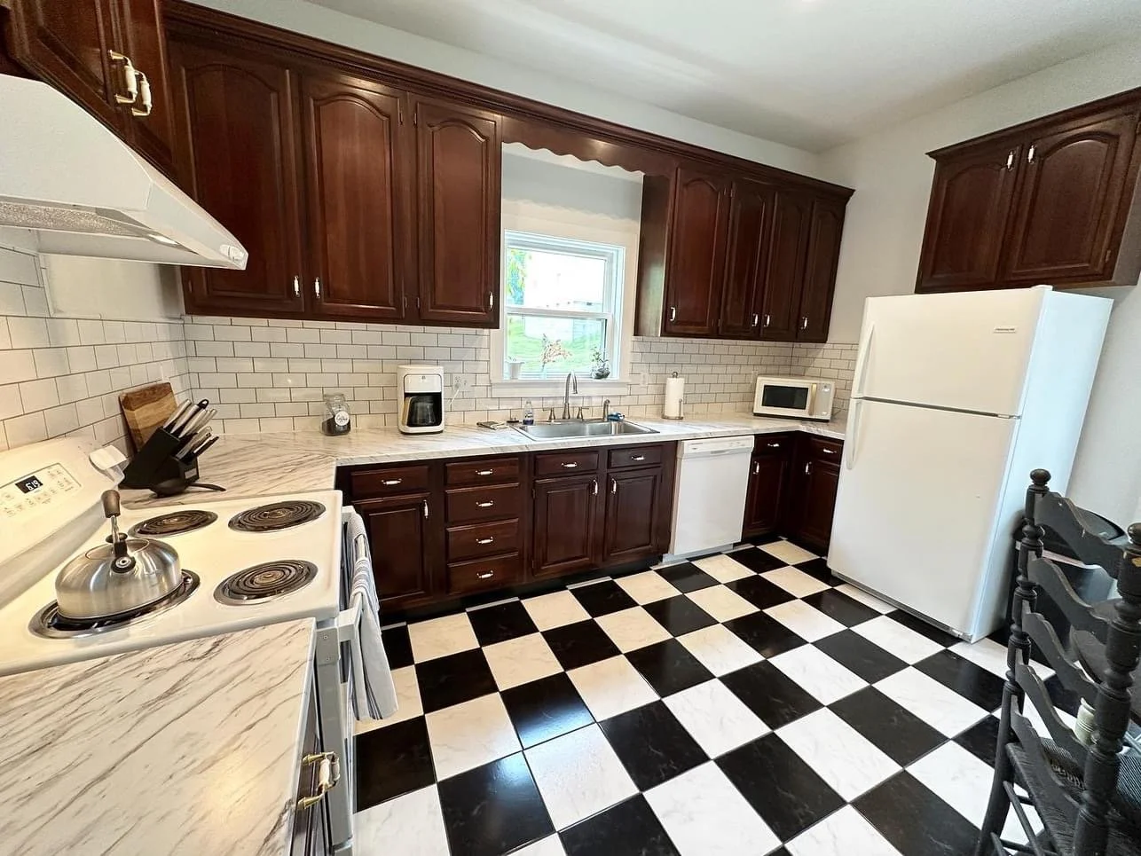 Kitchen with dark wood cabinets, white appliances, checkered black and white floor, white subway tile backsplash, window above the sink, microwave, coffee maker, and knife block on the counter.
