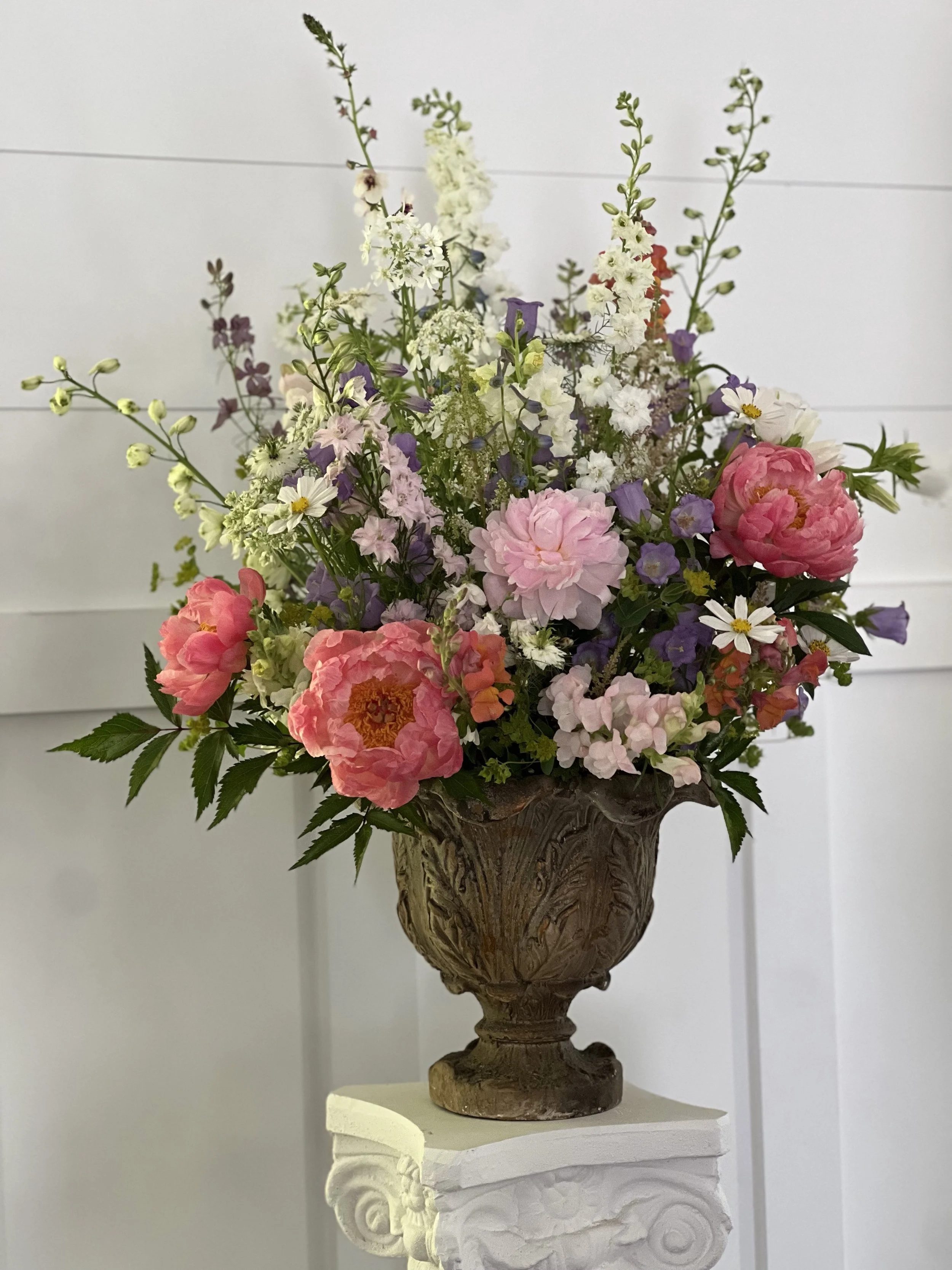 A tall floral arrangement featuring pink, white, purple, and green flowers in an ornate carved wooden vase on a white pedestal against a light-colored wall.