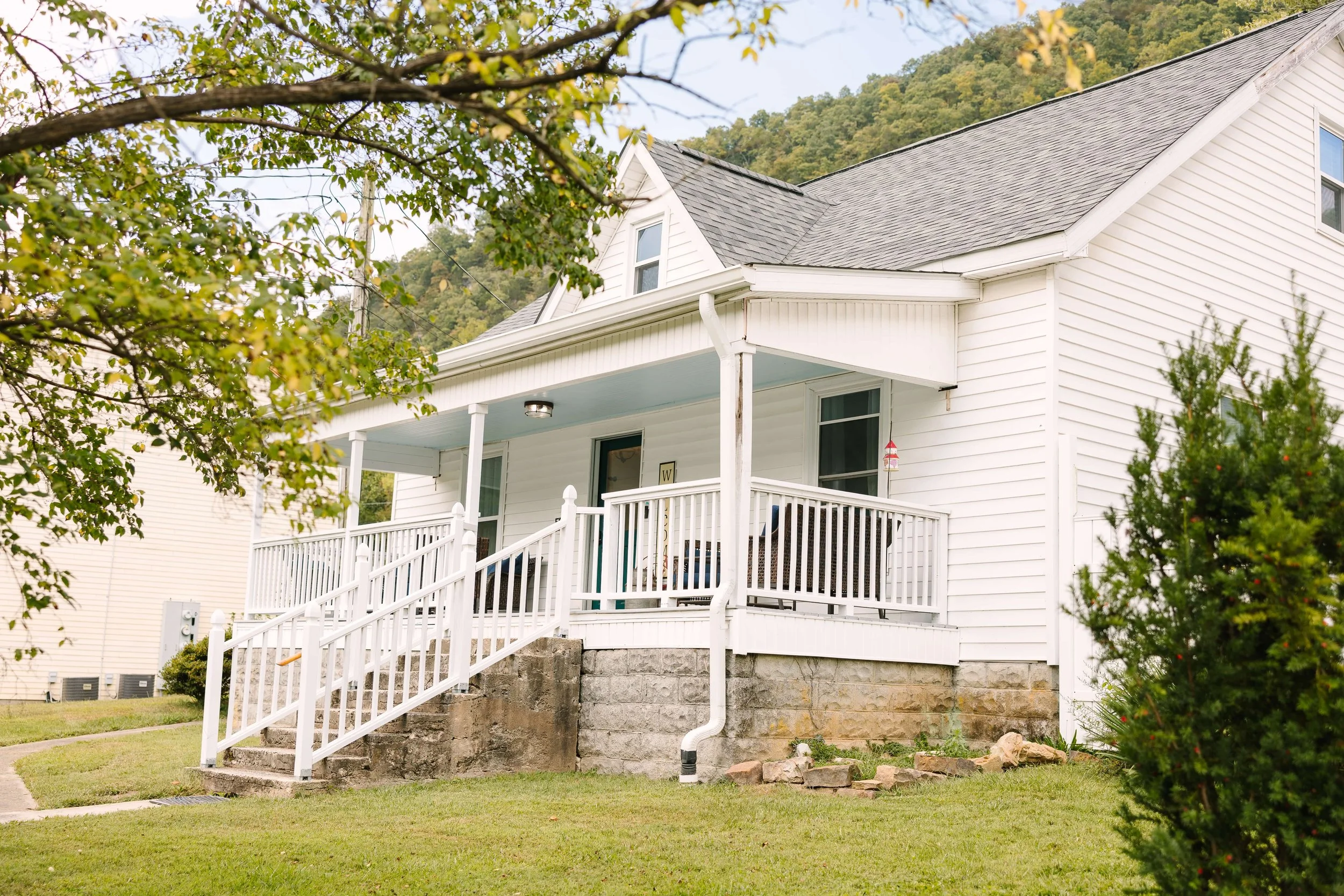 Front view of a white two-story house with a front porch, stairs, and railing, surrounded by green grass, trees, and hills in the background.
