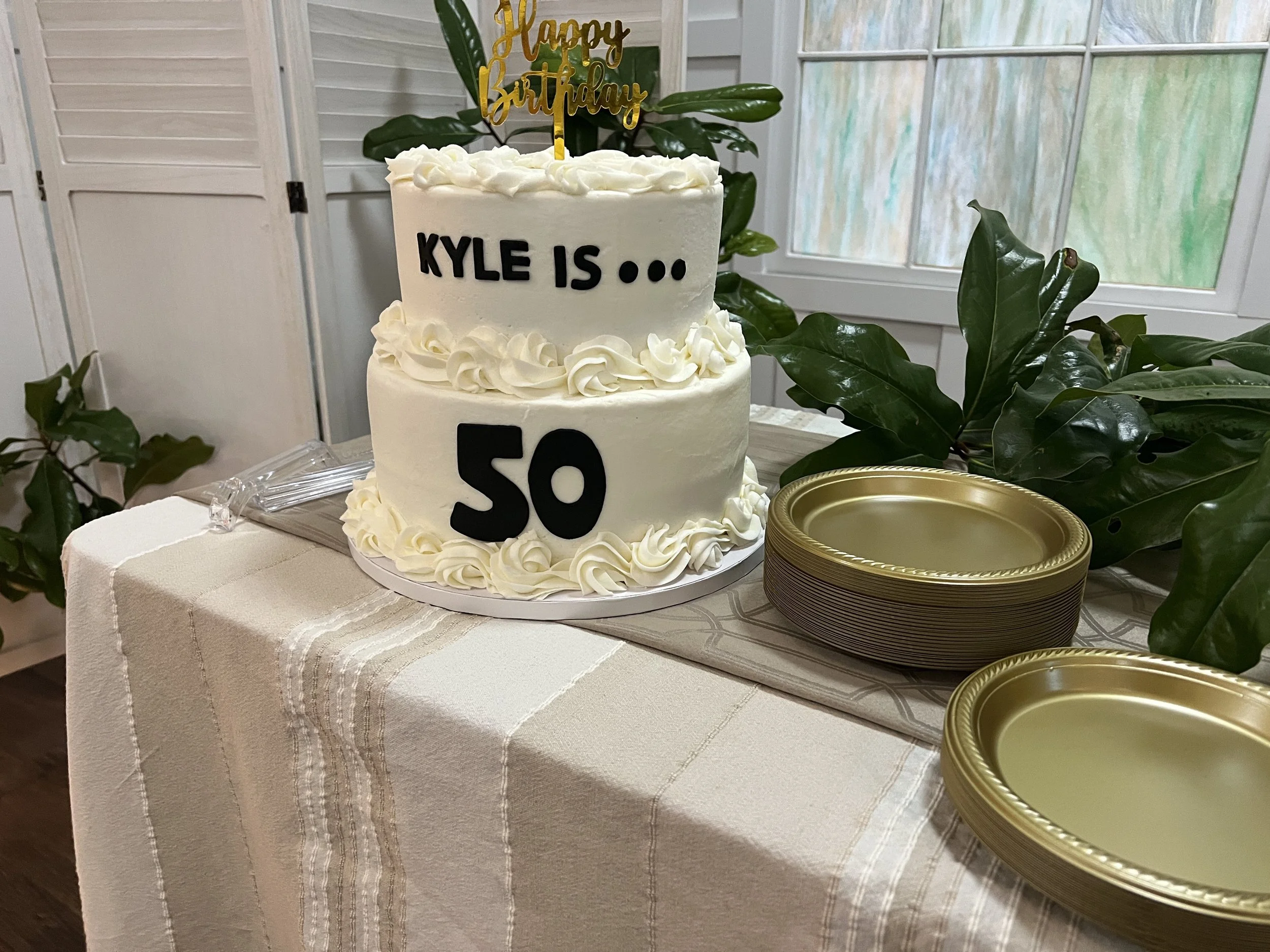A two-tiered white birthday cake with piped icing decorations. The top tier has a gold "Happy Birthday" cake topper, and the side of the cake has black letters spelling out "KYLE IS ..." and a large black number "50" below. The cake is on a white tab