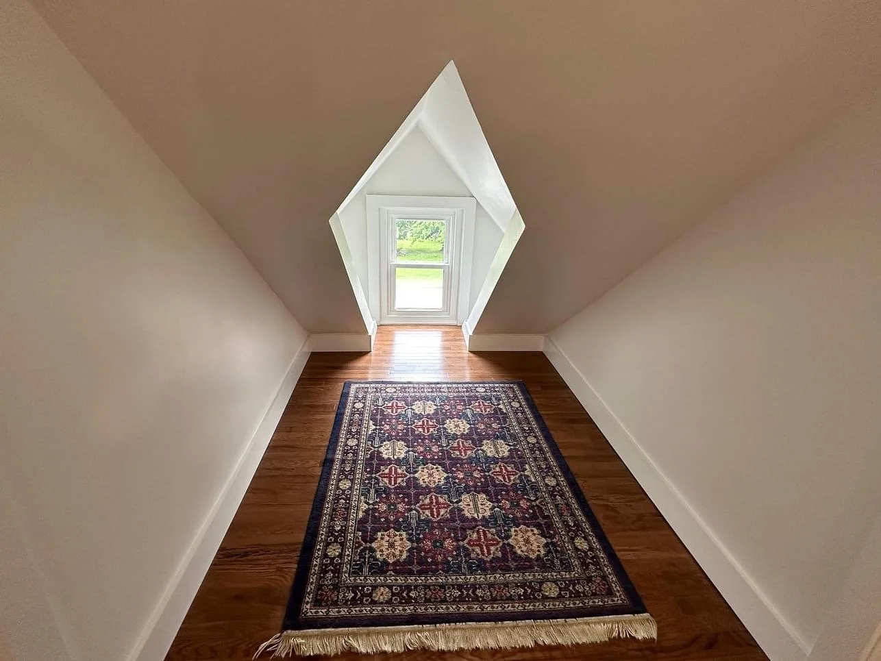 Small room with wood floor, a colorful rug, sloped ceiling, and a window with a view of green grass outside.