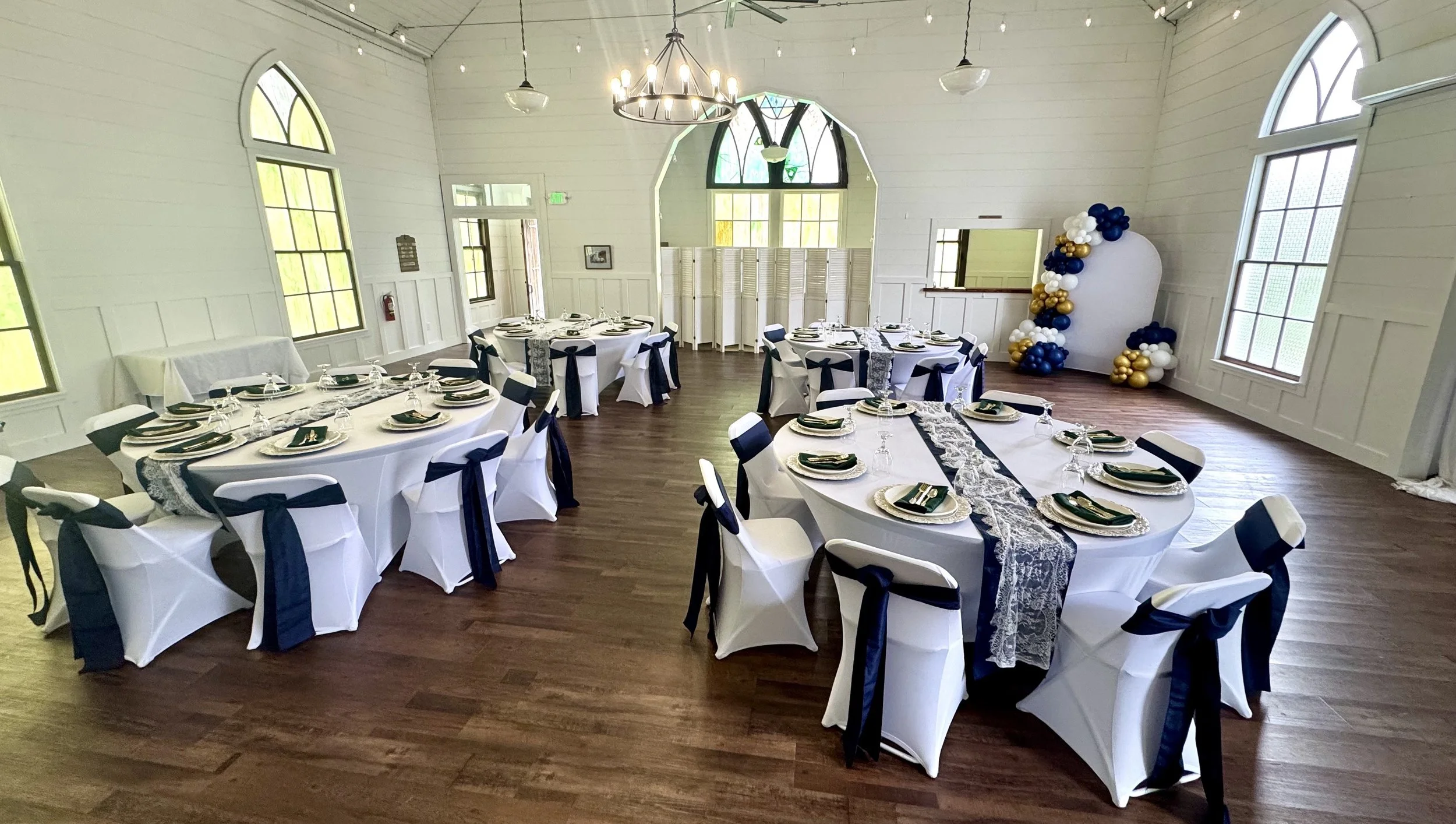 A decorated event room with round tables covered in white tablecloths, set with plates, napkins, and glassware for a formal gathering, featuring black and white chair covers with bow ties, large arched windows, wooden floors, and a balloon decoration in the corner.