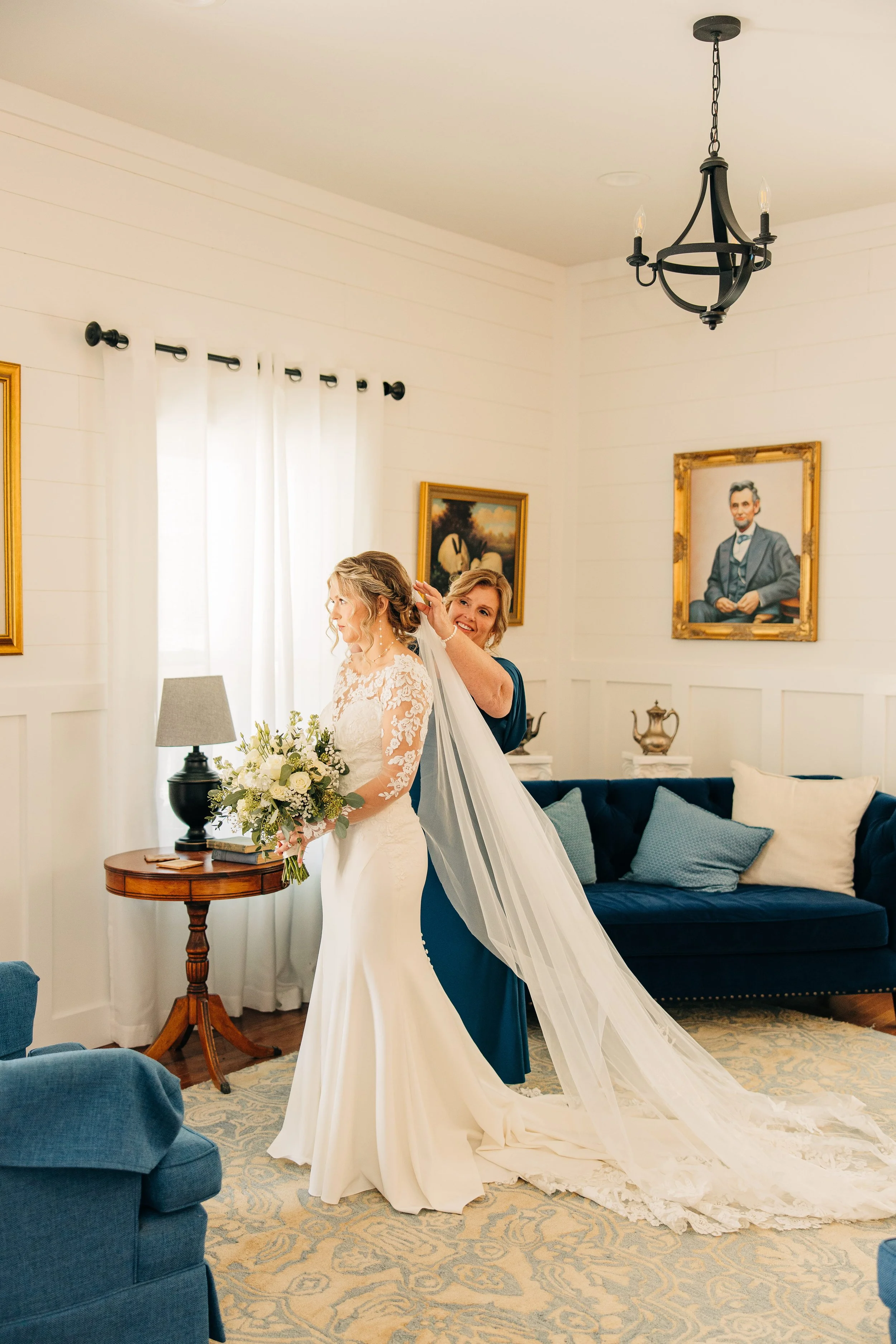 A woman in a white wedding dress holding a bouquet of white flowers, being assisted by another woman, in a room with cream-colored walls, paintings, a chandelier, and blue furniture.