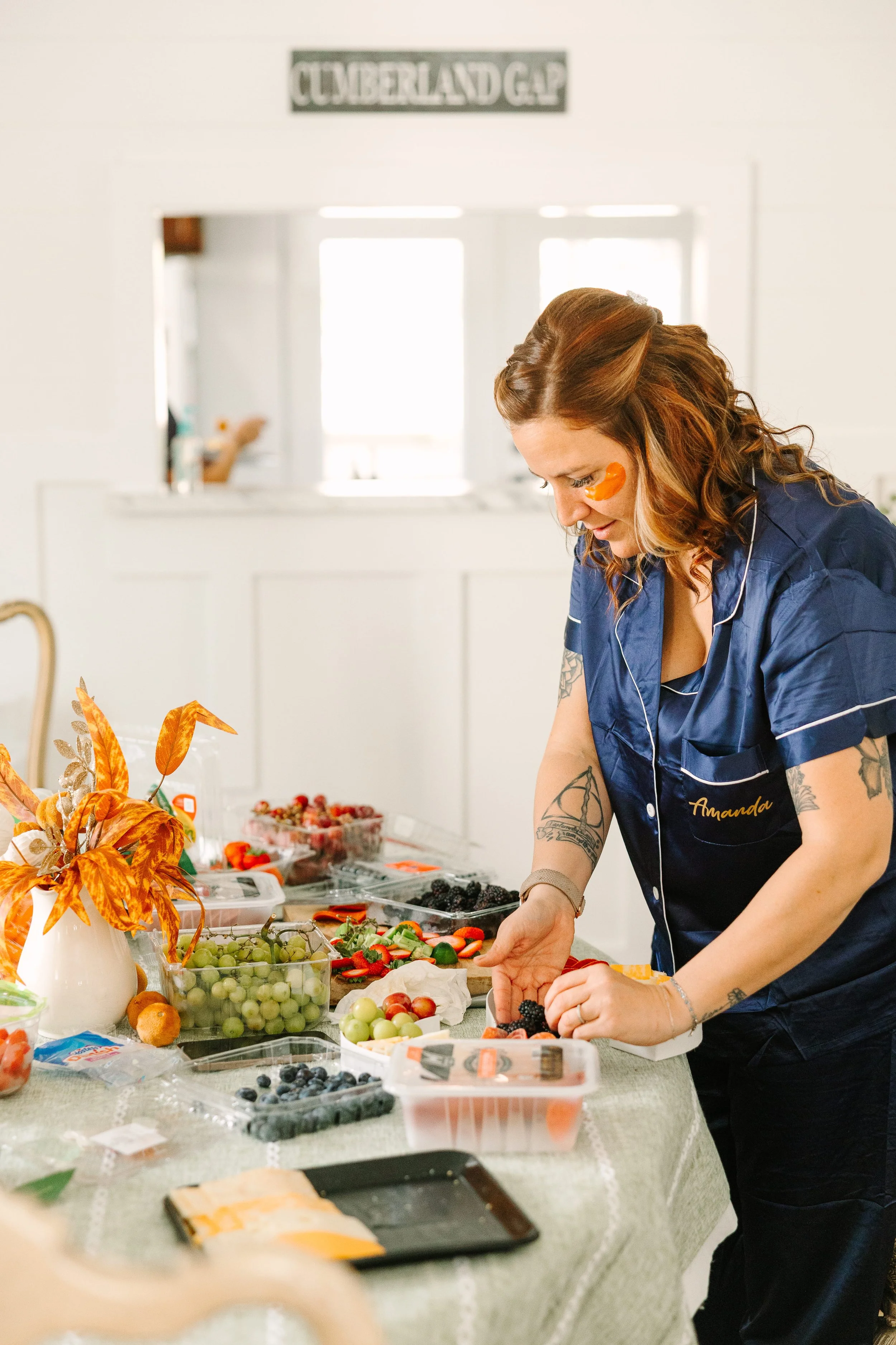 Woman in blue work shirt with name tag 'Amanda' arranging strawberries and other fruits on a buffet table, surrounded by grapes, blueberries, vegetables, and decorative fall-themed items in a bright, home-like kitchen setting.