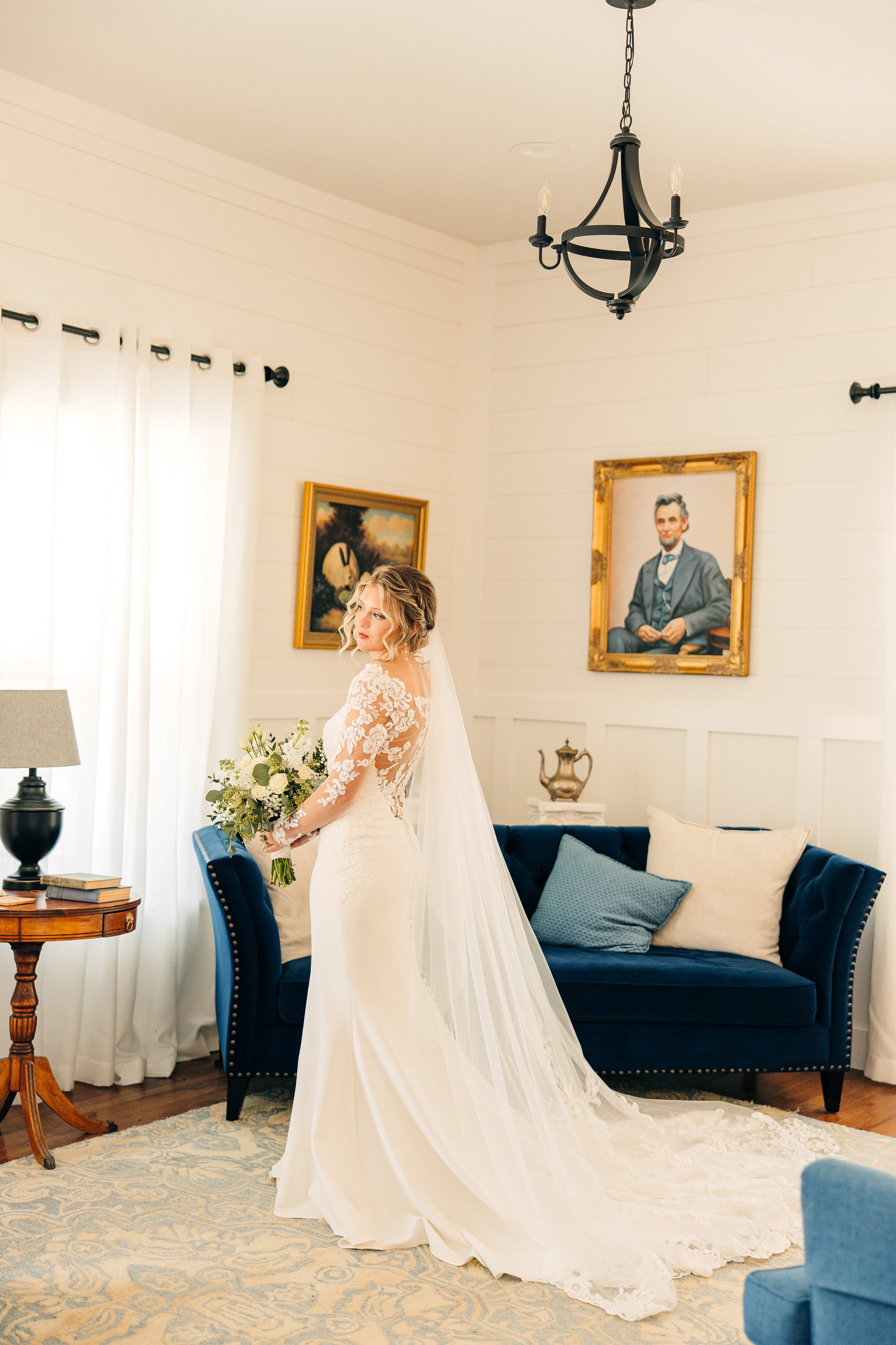 A bride in a white wedding dress with lace sleeves holding a bouquet of flowers in a living room with a blue sofa and framed paintings on the wall.