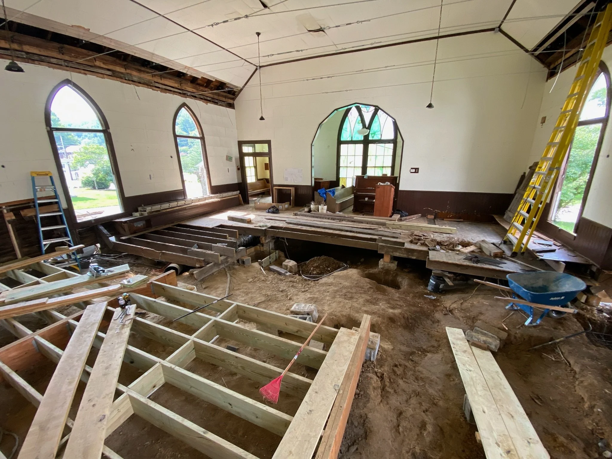 Interior of a building under renovation with exposed ceiling and framing, ladders, construction tools, and large windows allowing natural light.