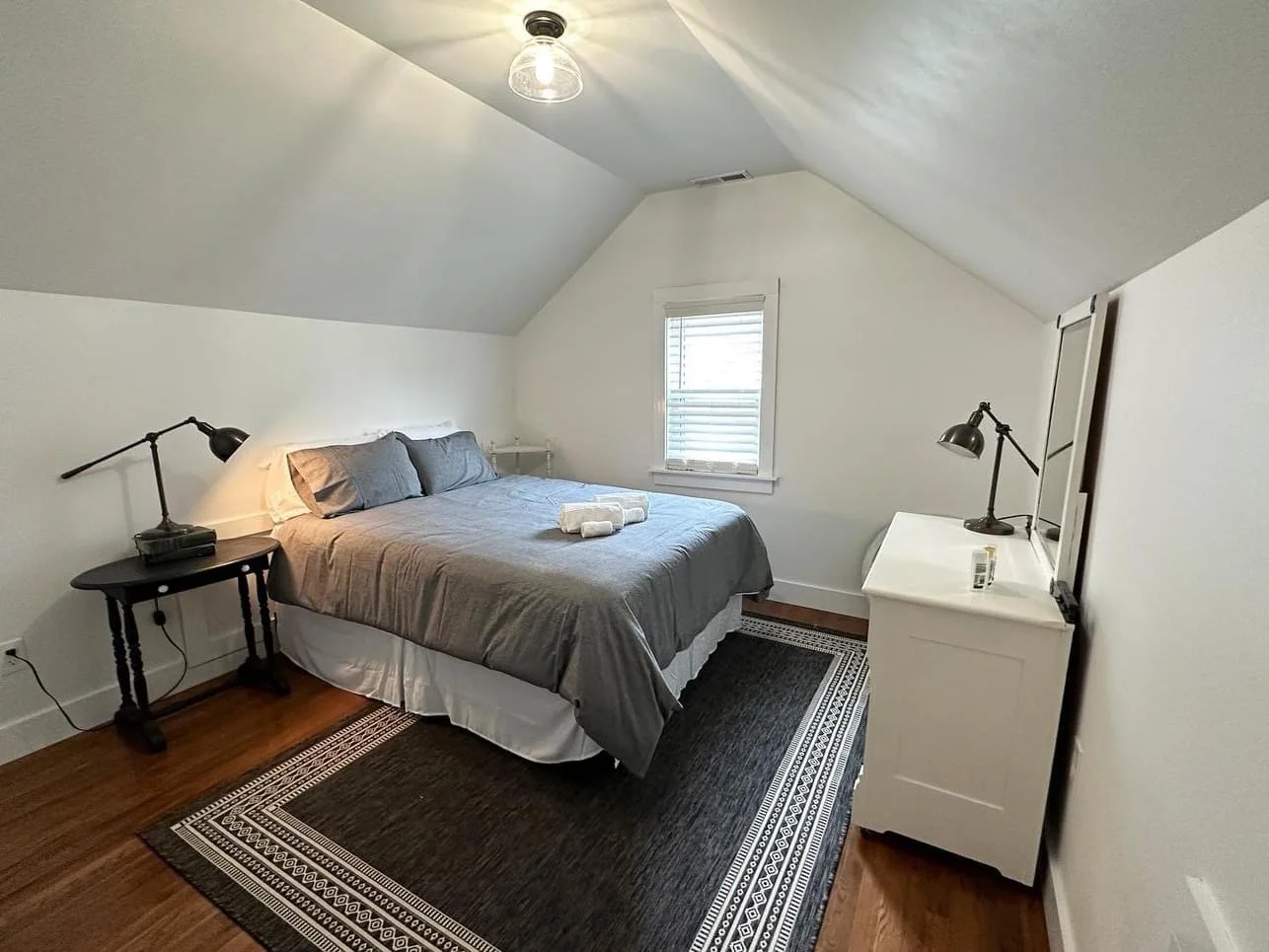 Cozy bedroom with a sloped ceiling, white walls, a bed with gray bedding, two black lamps on either side, a small white dresser, and a window with blinds.