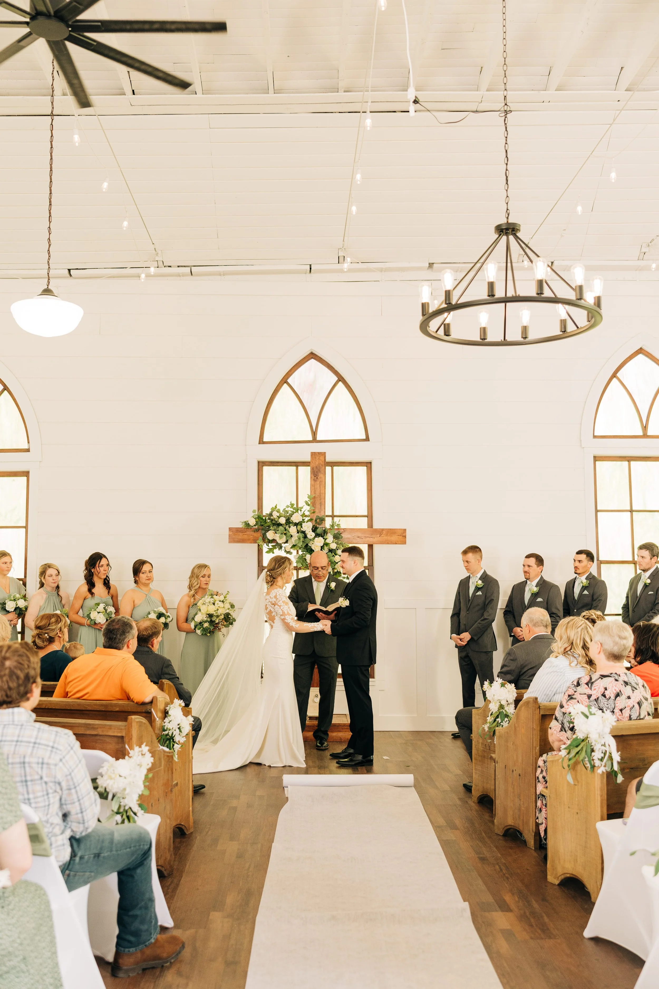 A wedding ceremony with a bride and groom standing before a pastor, with bridal party members and guests seated inside a white chapel with wooden accents and a cross decorated with flowers behind them.