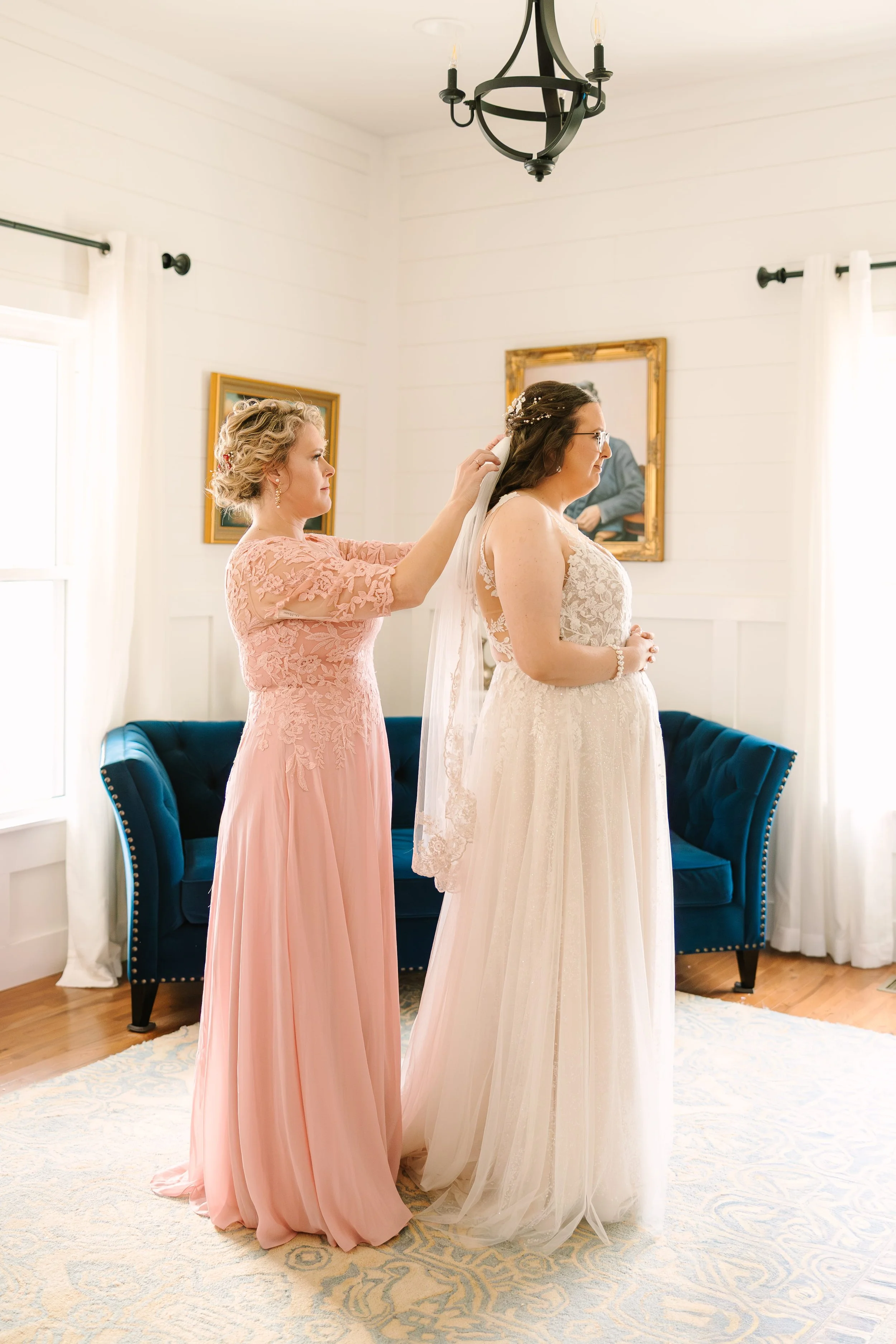 A bride in a lace wedding dress is getting ready for her wedding with the assistance of a woman in a pink dress, in a bright room with white walls, a blue couch, and framed pictures on the wall.
